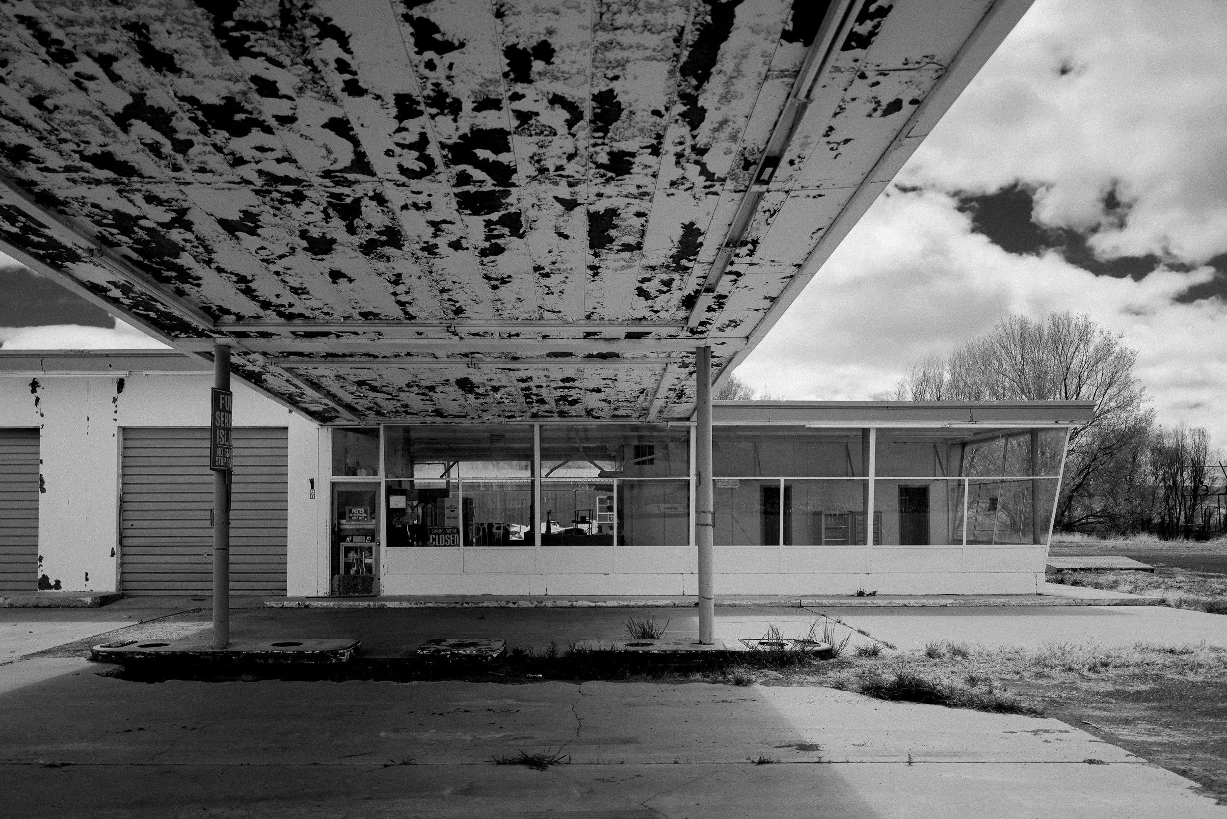 An abandoned gas station with a peeling cantilevered canopy roof and glass-walled office.