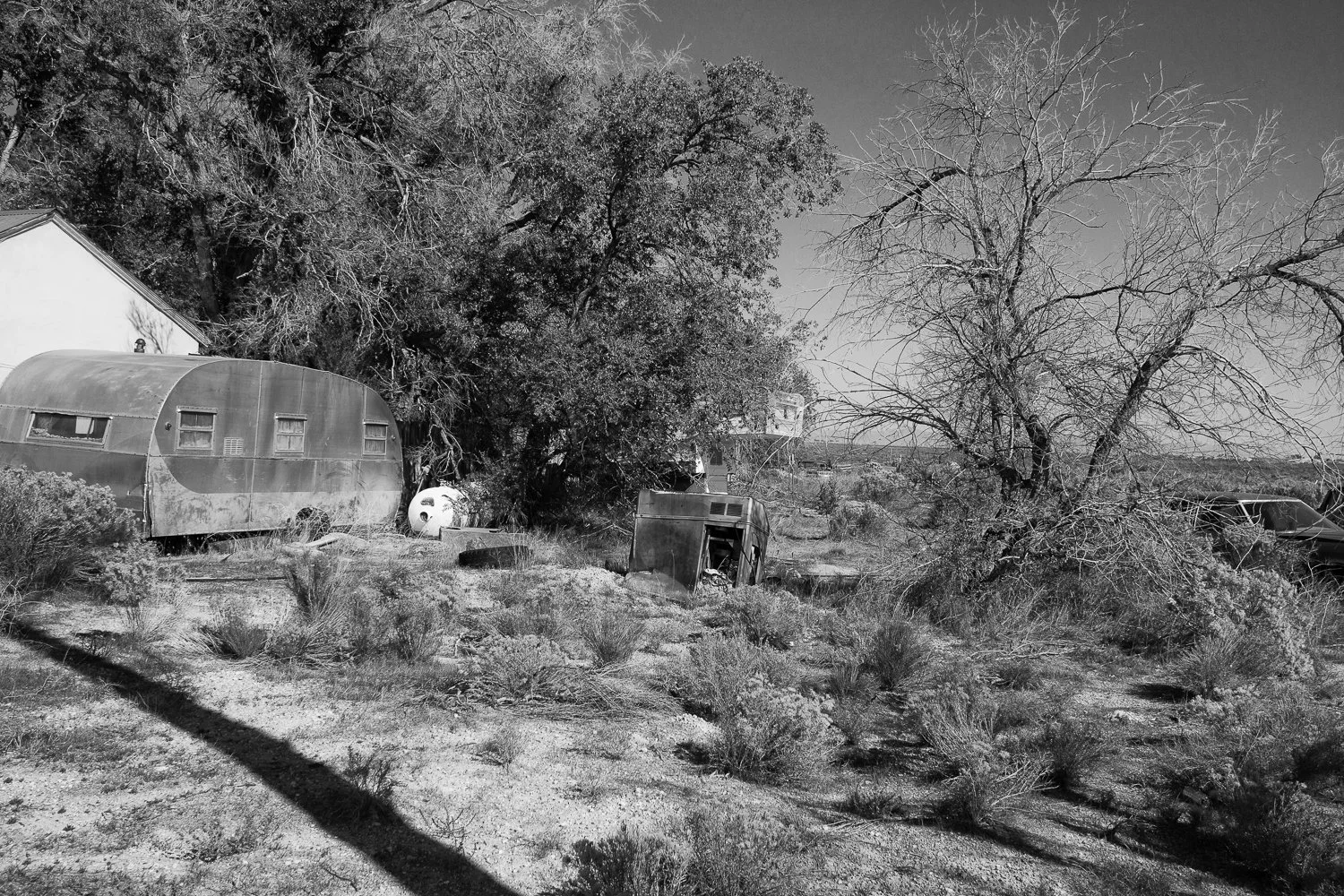 An abandoned vintage travel trailer and scattered debris beneath desert trees.
