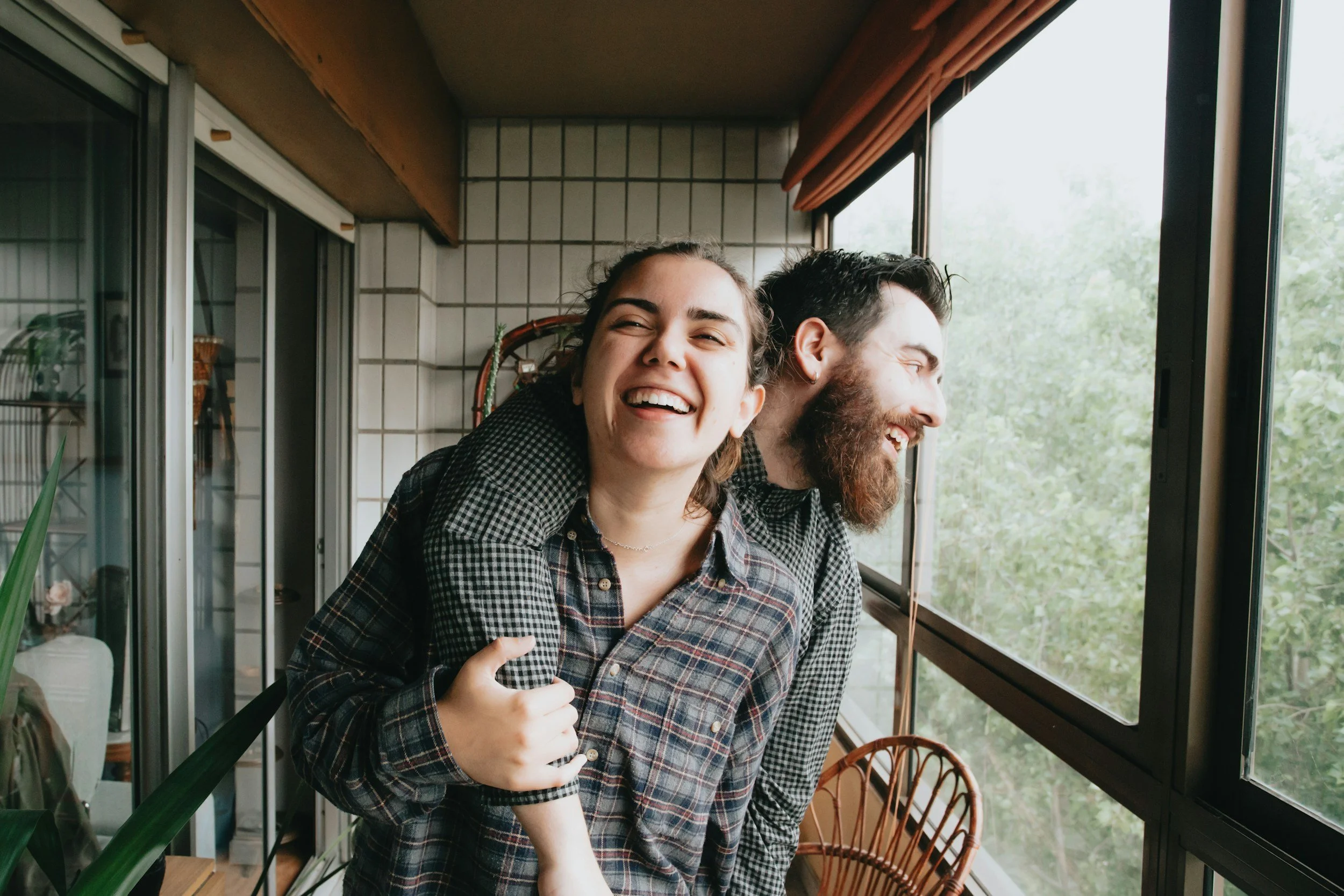 A woman giving a piggyback ride to a man, both smiling and looking outside a large window in a cozy indoor space with a green view outside.