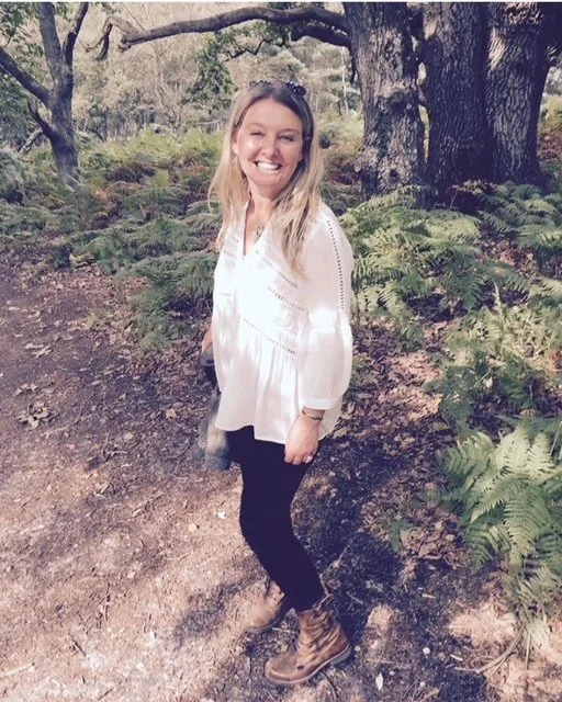 A smiling woman with blonde hair, wearing a white blouse, black pants, and hiking boots, standing in a forest with trees and green ferns.