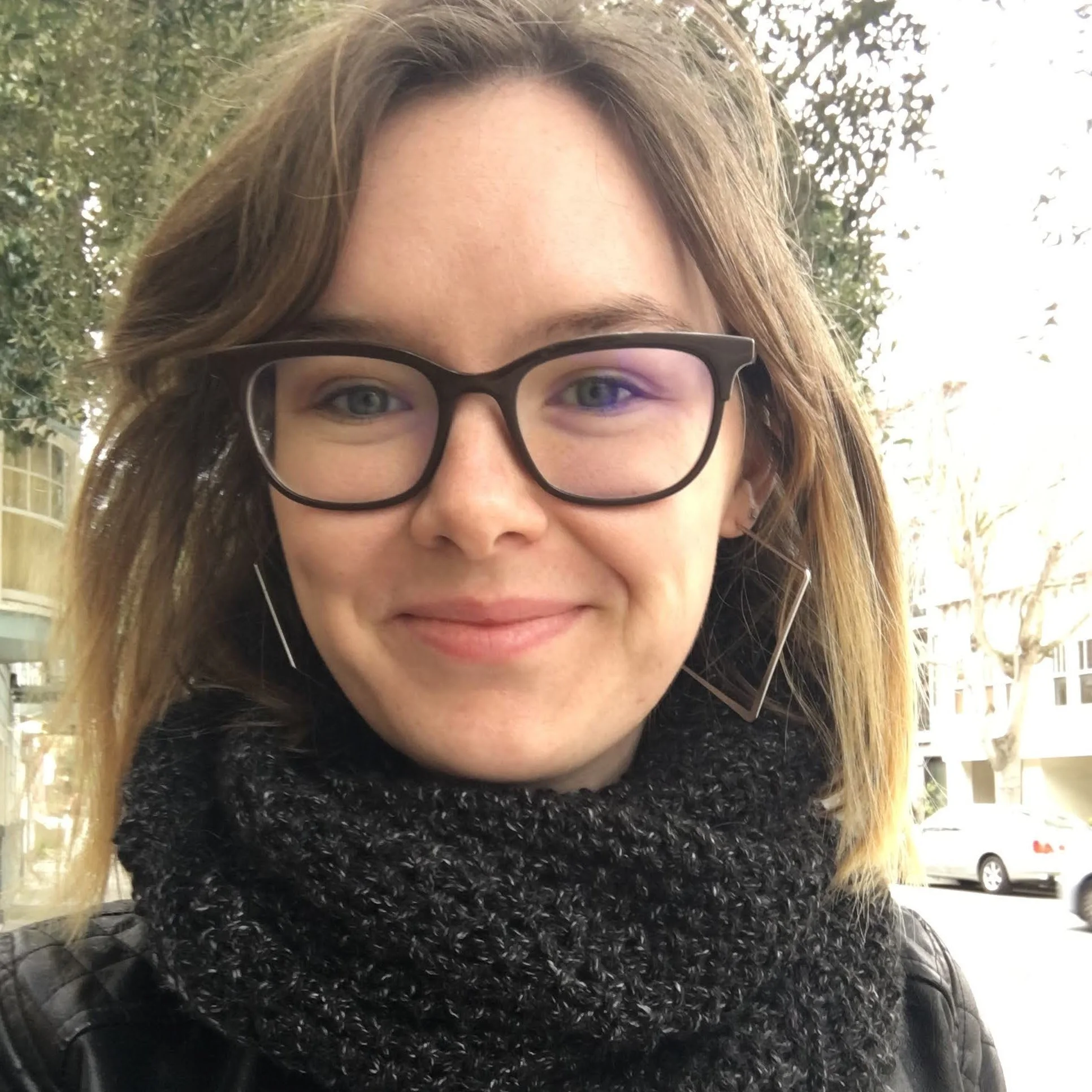 Rachael Salisbury smiling wearing dark rimmed glasses, hoop earrings, and a dark gray scarf