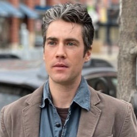 Connor Gibson looking off into the distance, wearing a light brown blazer and a denim chambray shirt, unbuttoned at the top.
