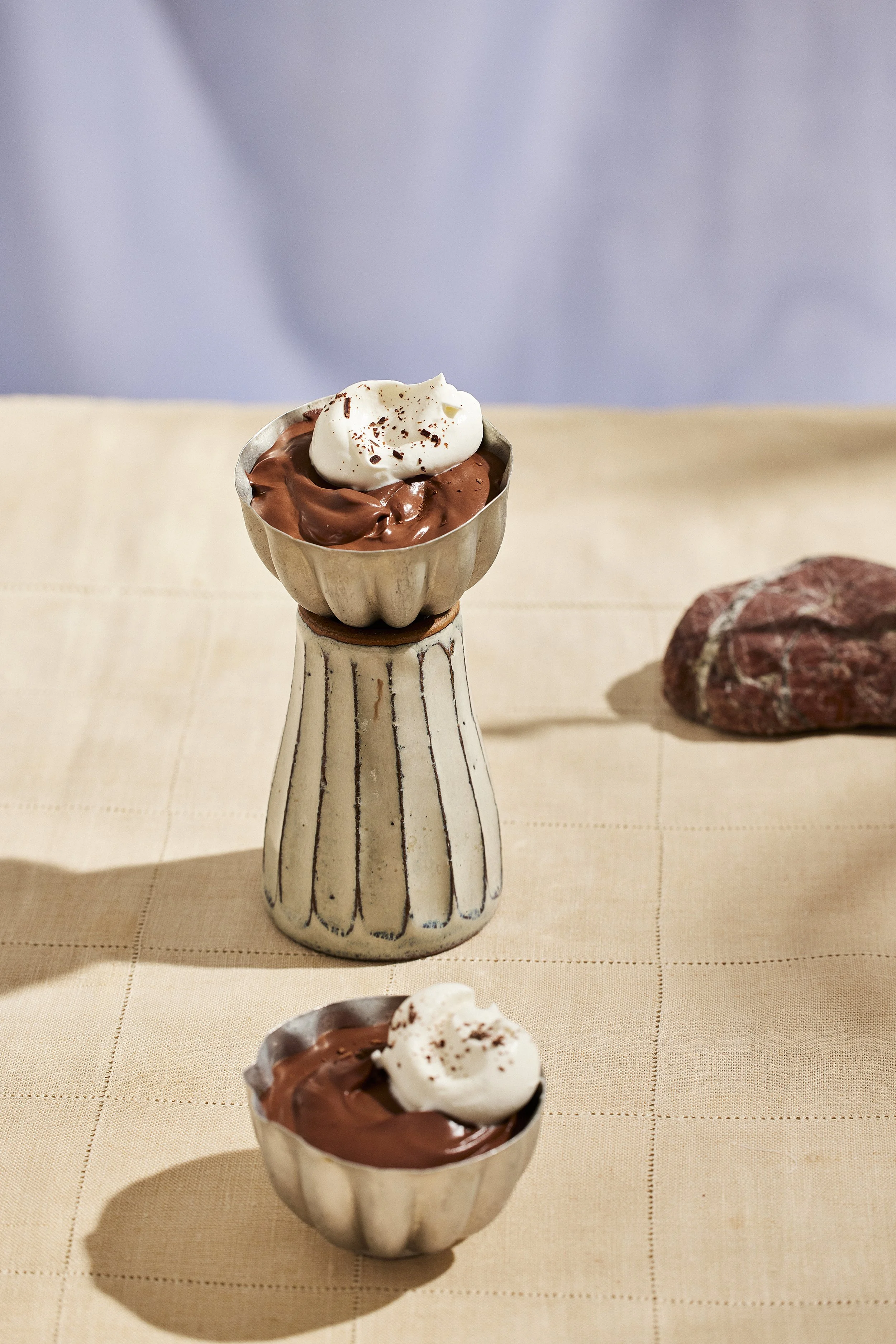 Two metal bowls filled with chocolate mousse topped with whipped cream on a beige tablecloth, with a stone in the background.