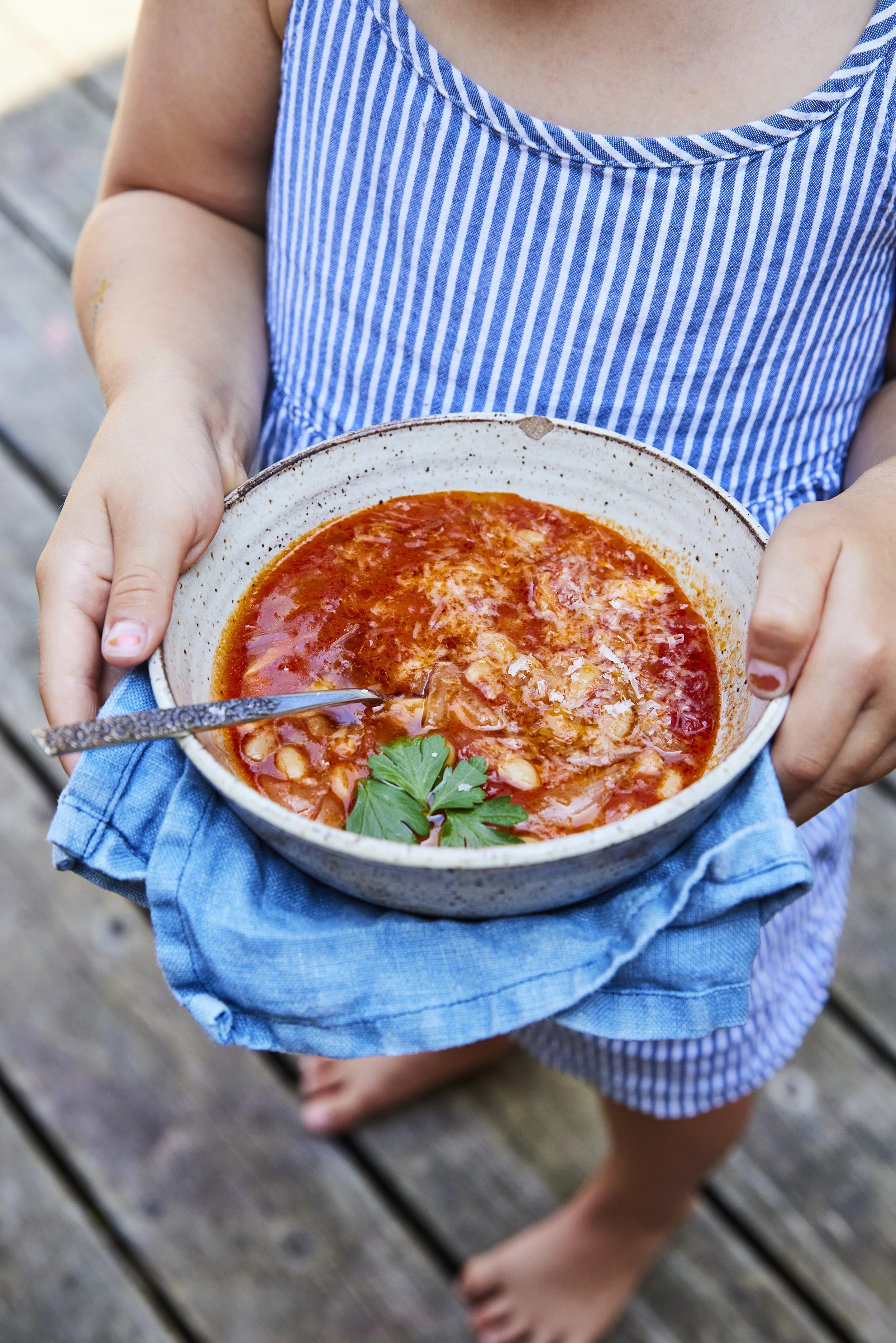 Child in a blue and white striped dress holding a bowl of red tomato soup garnished with cilantro, on a wooden deck.