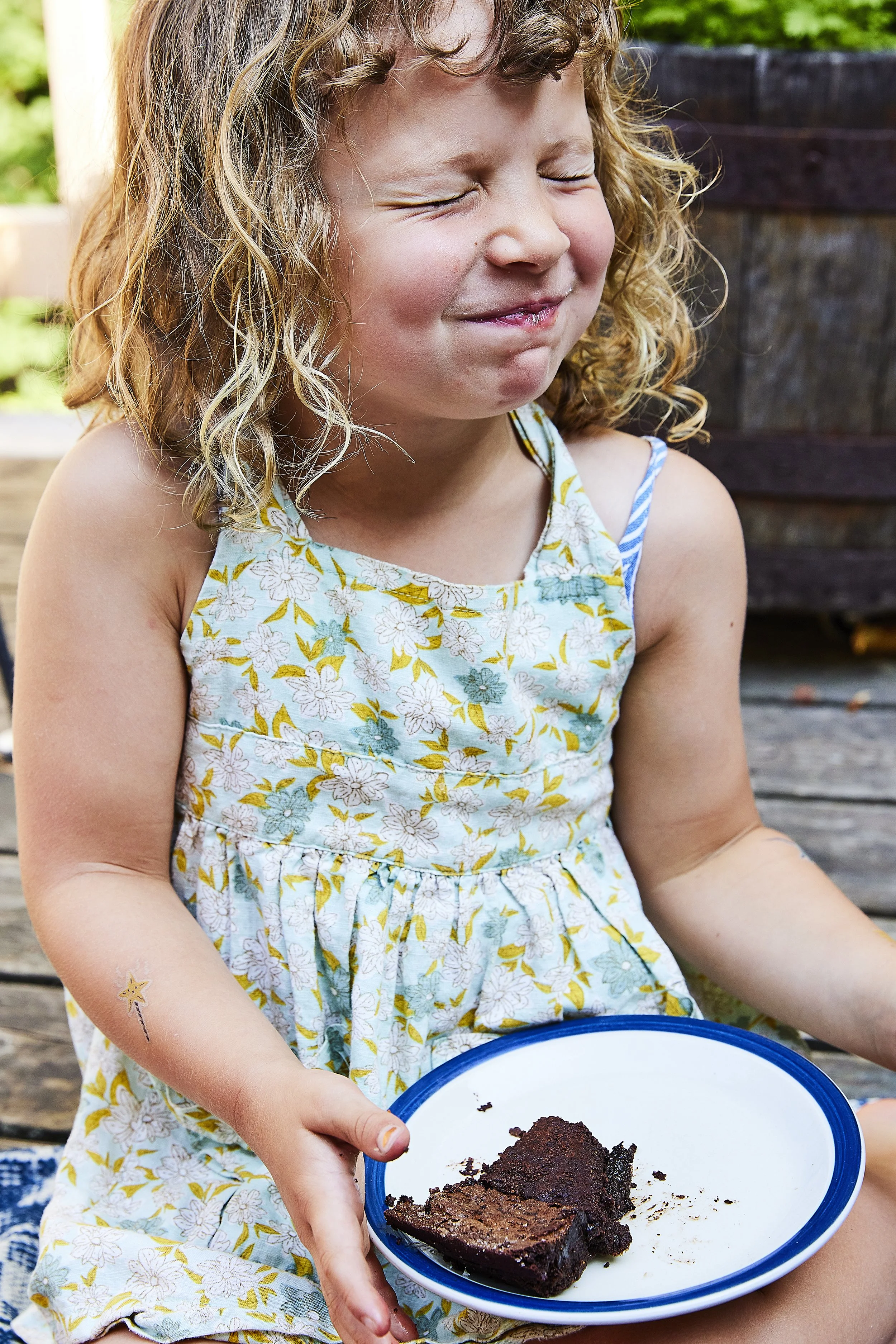 A young girl with curly blonde hair wearing a floral dress, holding a plate with a partially eaten chocolate cake, her eyes closed and a satisfied expression on her face.
