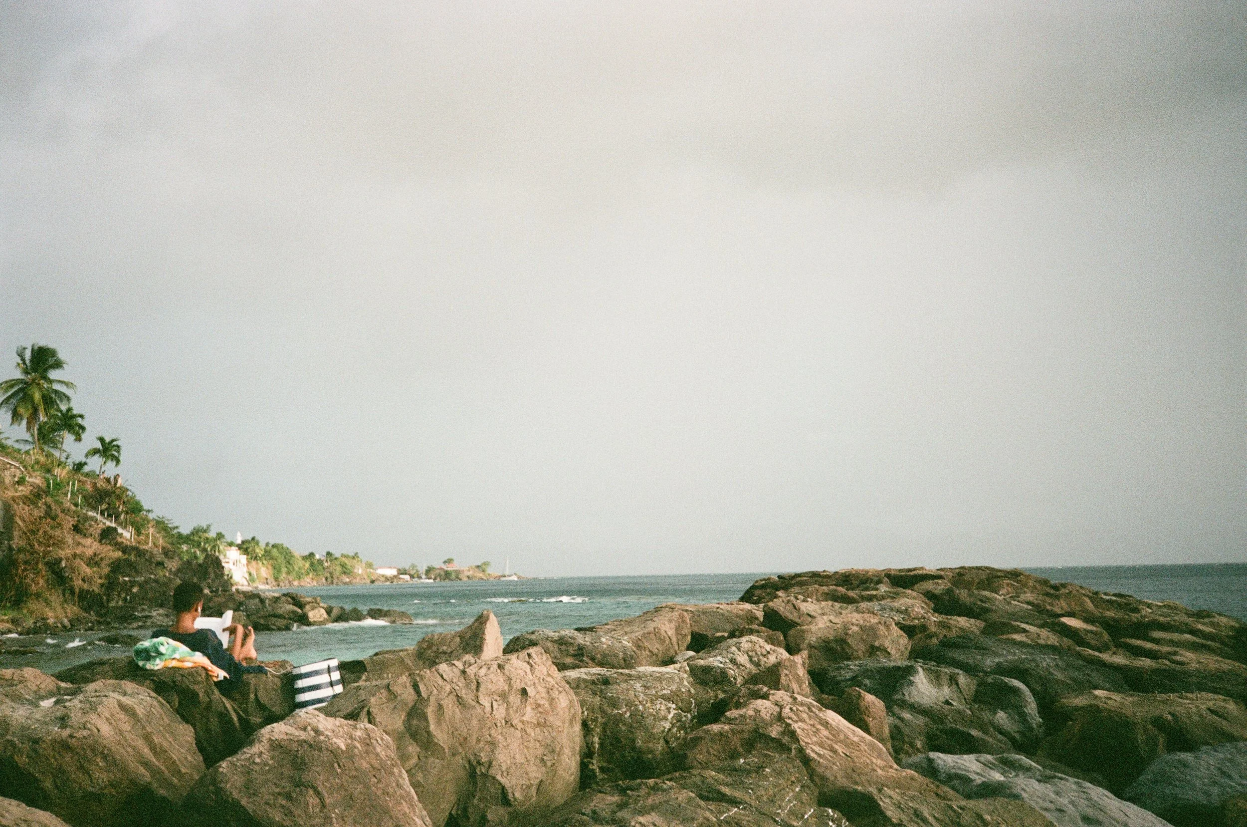 Person sitting on rocks by the ocean, reading a book, with palm trees and houses on a hillside in the background.