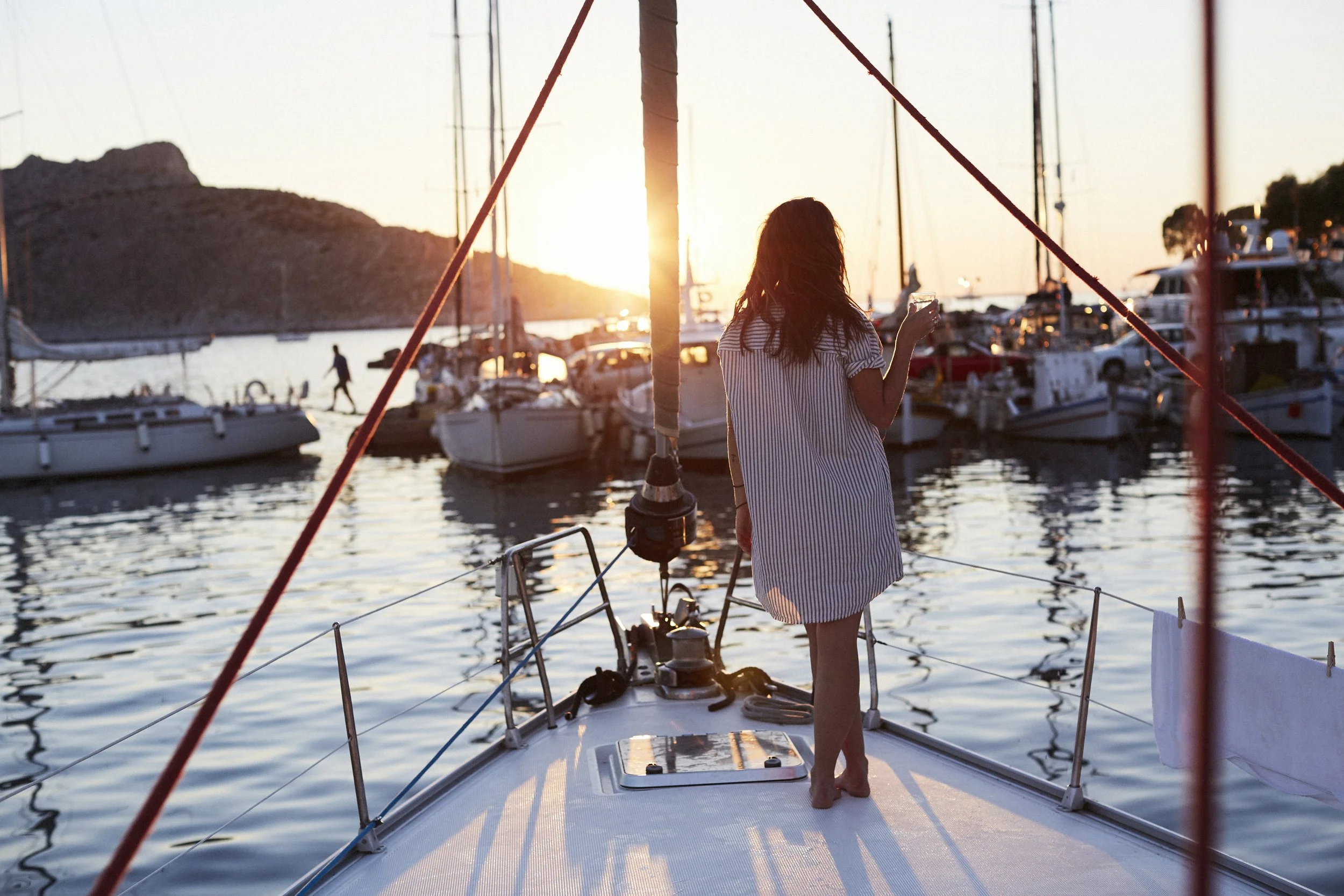 Woman standing on a boat at sunset, looking at the marina filled with docked sailboats and yachts, holding a drink in her hand.