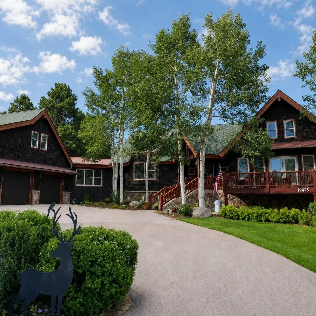 A two-story gray house with a front porch, surrounded by a garden with potted plants and flowers, and tall pine trees in the yard.