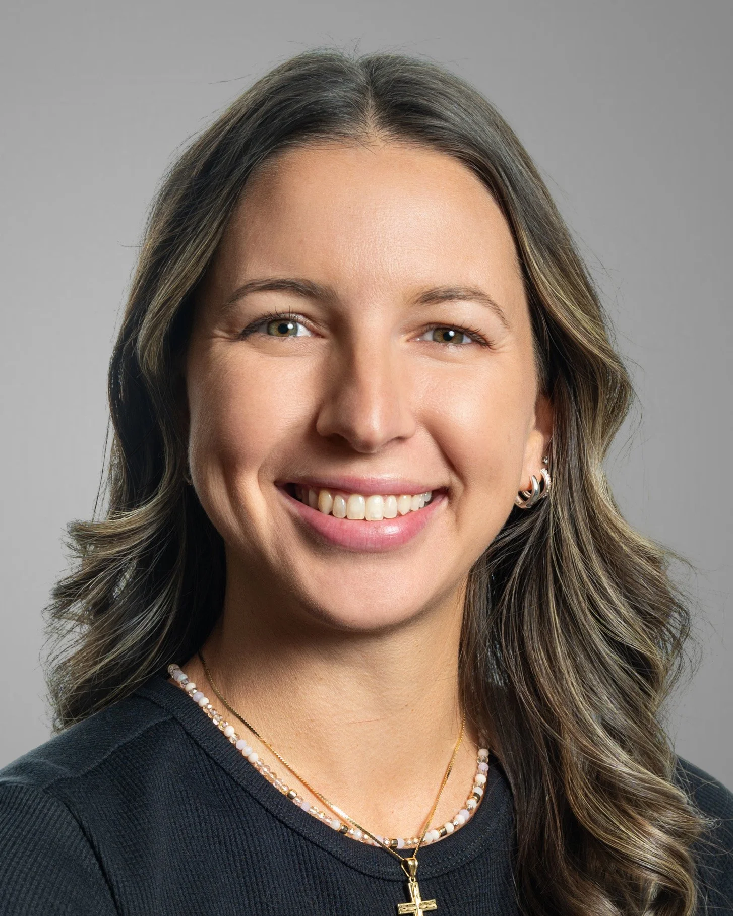 Smiling woman with long dark hair, wearing a green top, gold hoop earrings, and a necklace, on a white background.