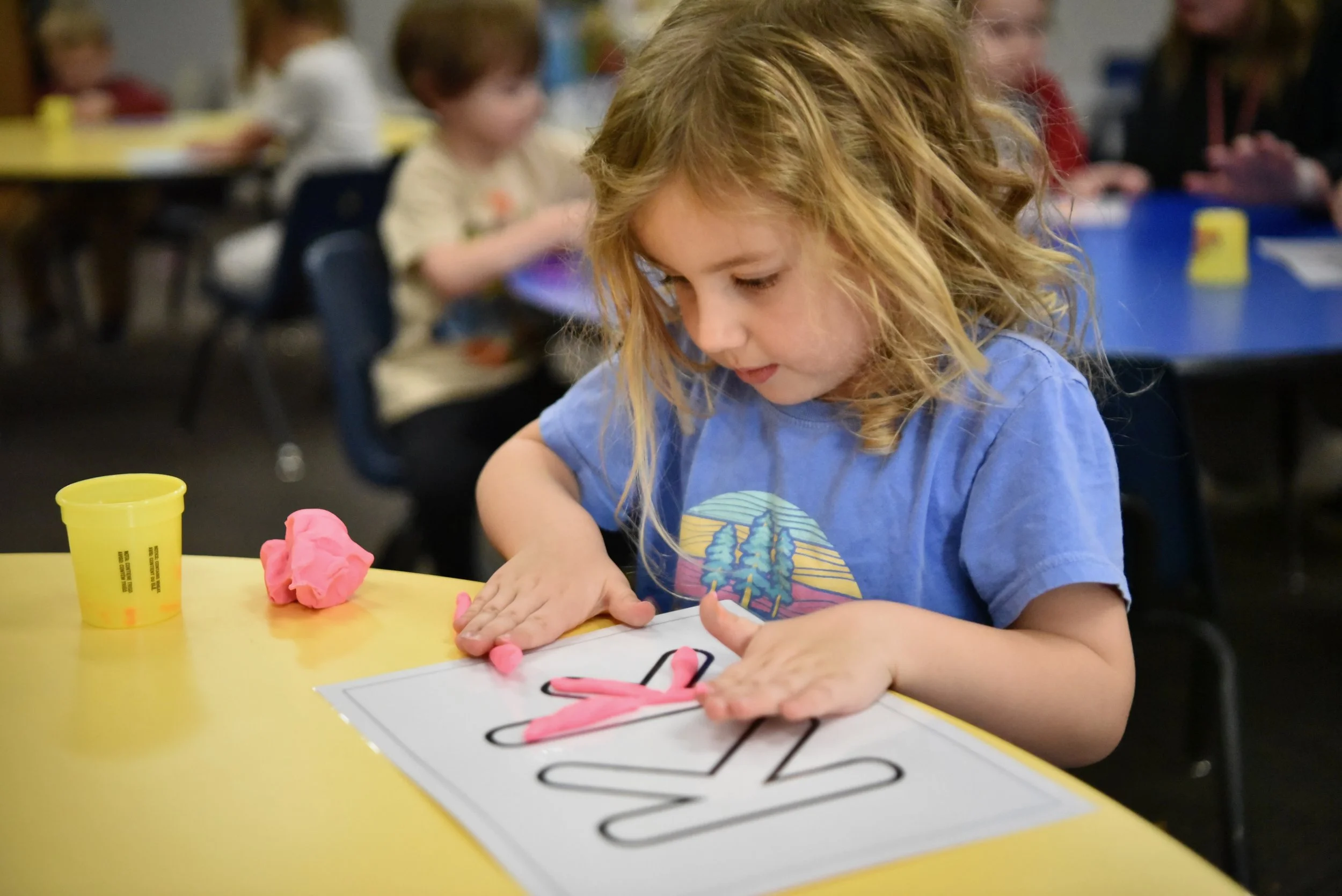 Child sitting at a school desk, hands clasped, with a toy bear in foreground.
