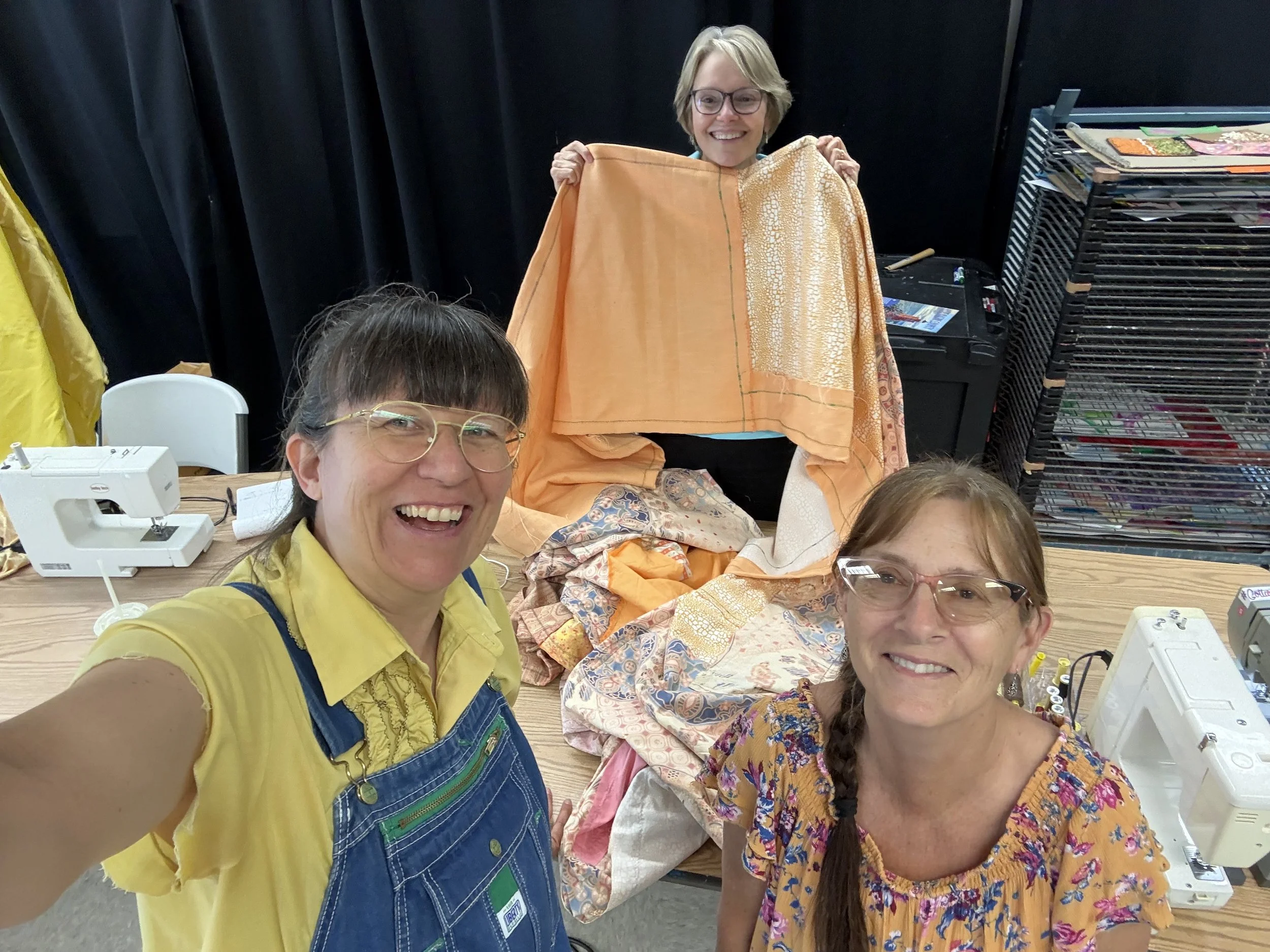 Three people smiling. One person holding up orange fabric. two sewing machines are on the table.