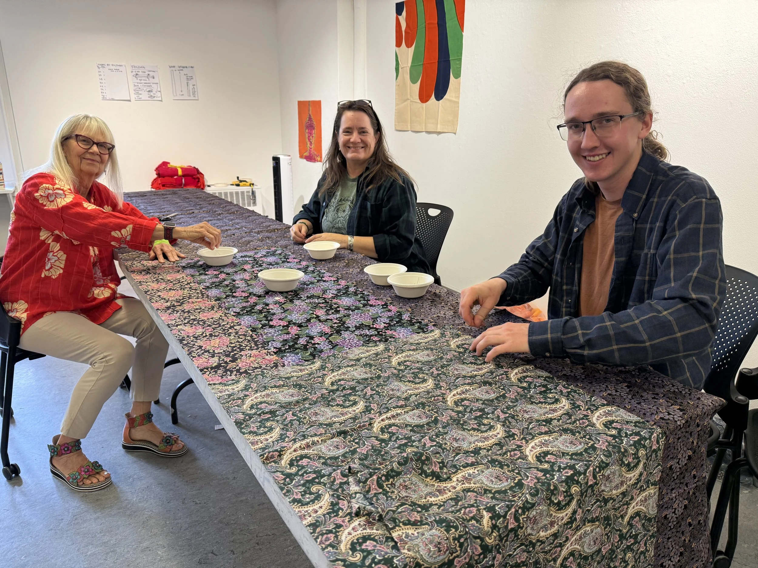 three people sitting at a table with fabric on it that they are pinning. all smiling.