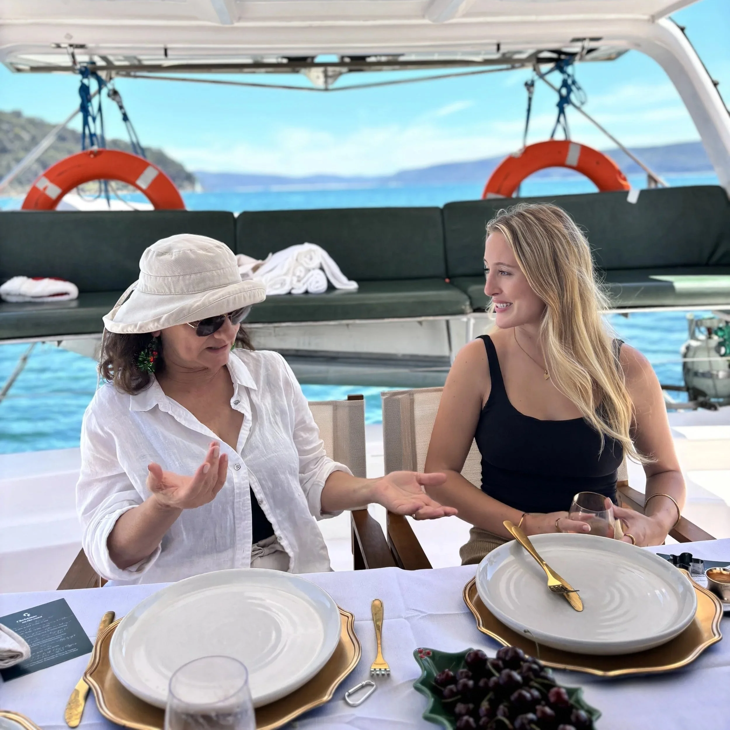 A table set for a meal on a boat with white dinner plates, gold-colored utensils, and a bowl of grapes. Two women are sitting at the table, one wearing a white shirt and a sun hat, the other with long blonde hair and a black tank top. The background shows the blue ocean and a coastline with hills.