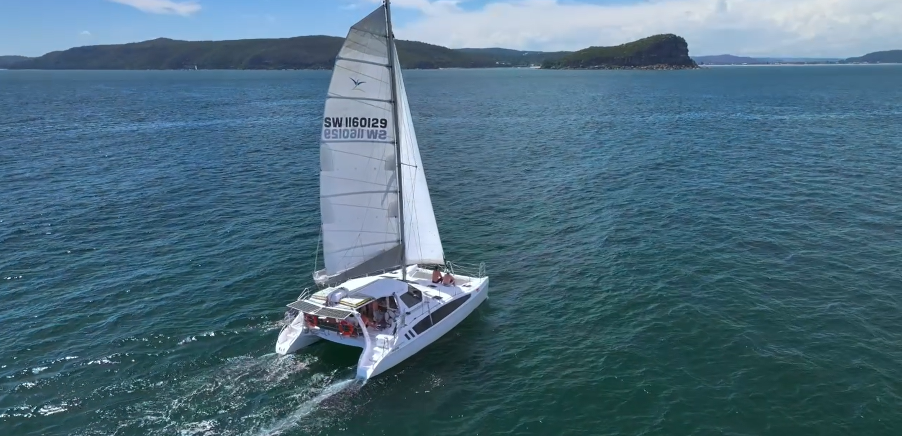 A white sailing catamaran with sails in the water near a coastline with hills and cliffs, under a partly cloudy sky.
