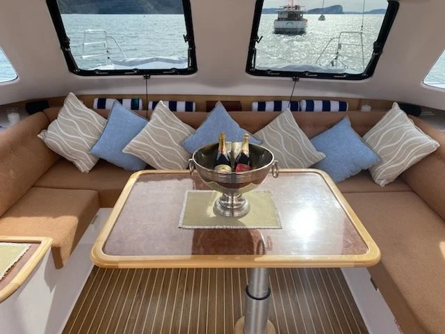 Interior of a boat with a wooden table, a metal ice bucket holding two bottles of champagne, and cushions on a U-shaped beige seating area. Windows show water and boats outside.
