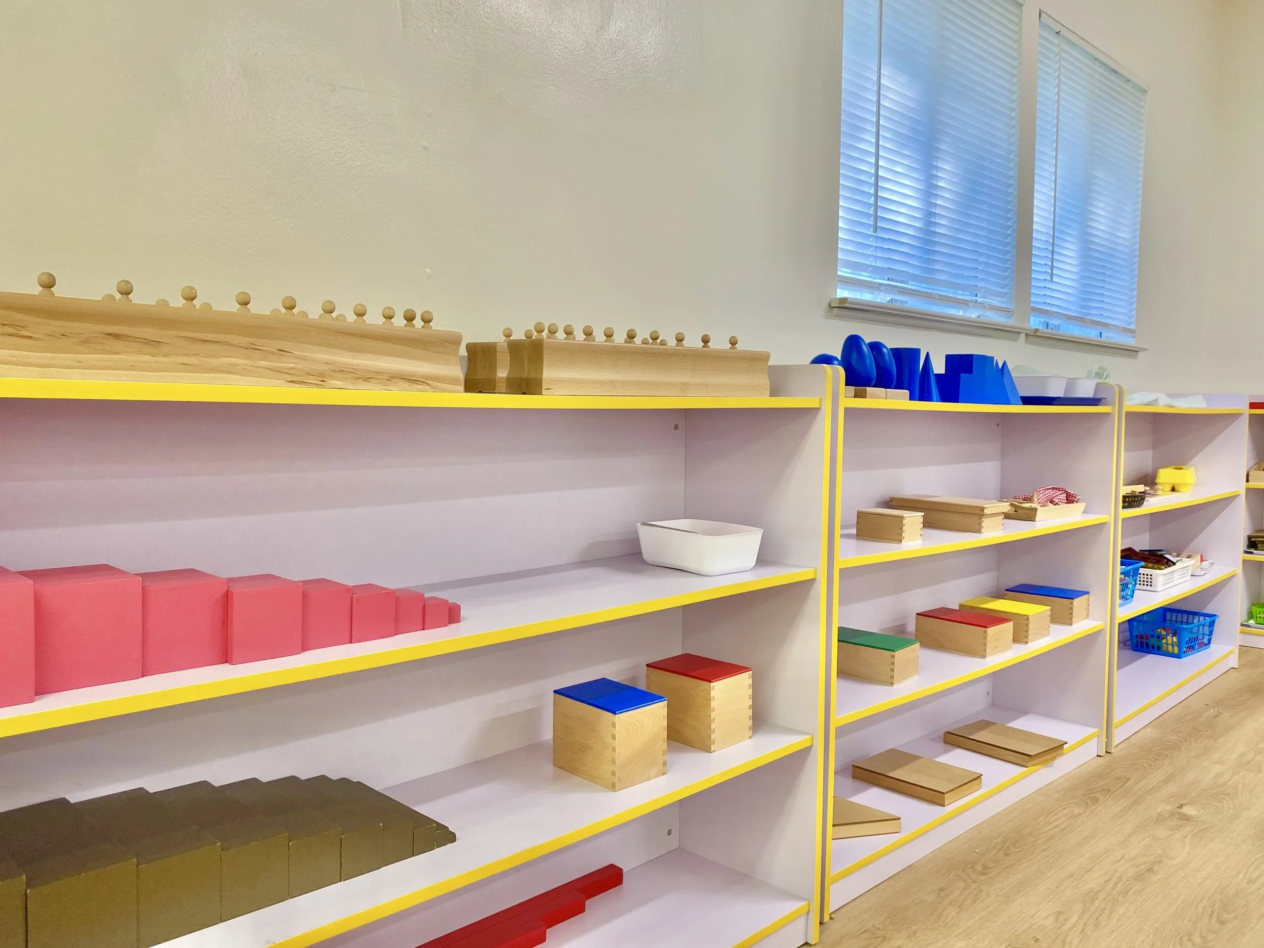 Colorful wooden Montessori shelf with geometric blocks, shapes, and toys in a classroom setting with large windows and white blinds.