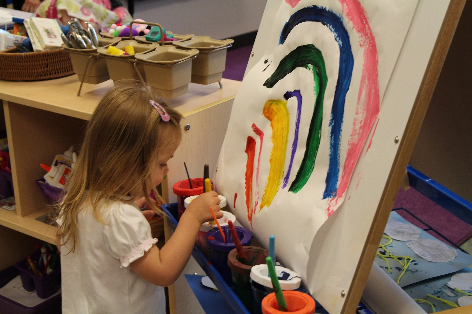 A young girl painting a rainbow on a large sheet of paper with colorful paints and brushes.