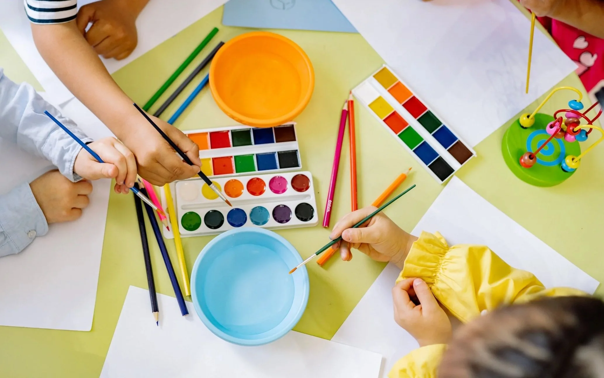 Children's hands engaging in watercolor painting and craft activities on a table along with colorful supplies, including paints, brushes, paper, and small containers.
