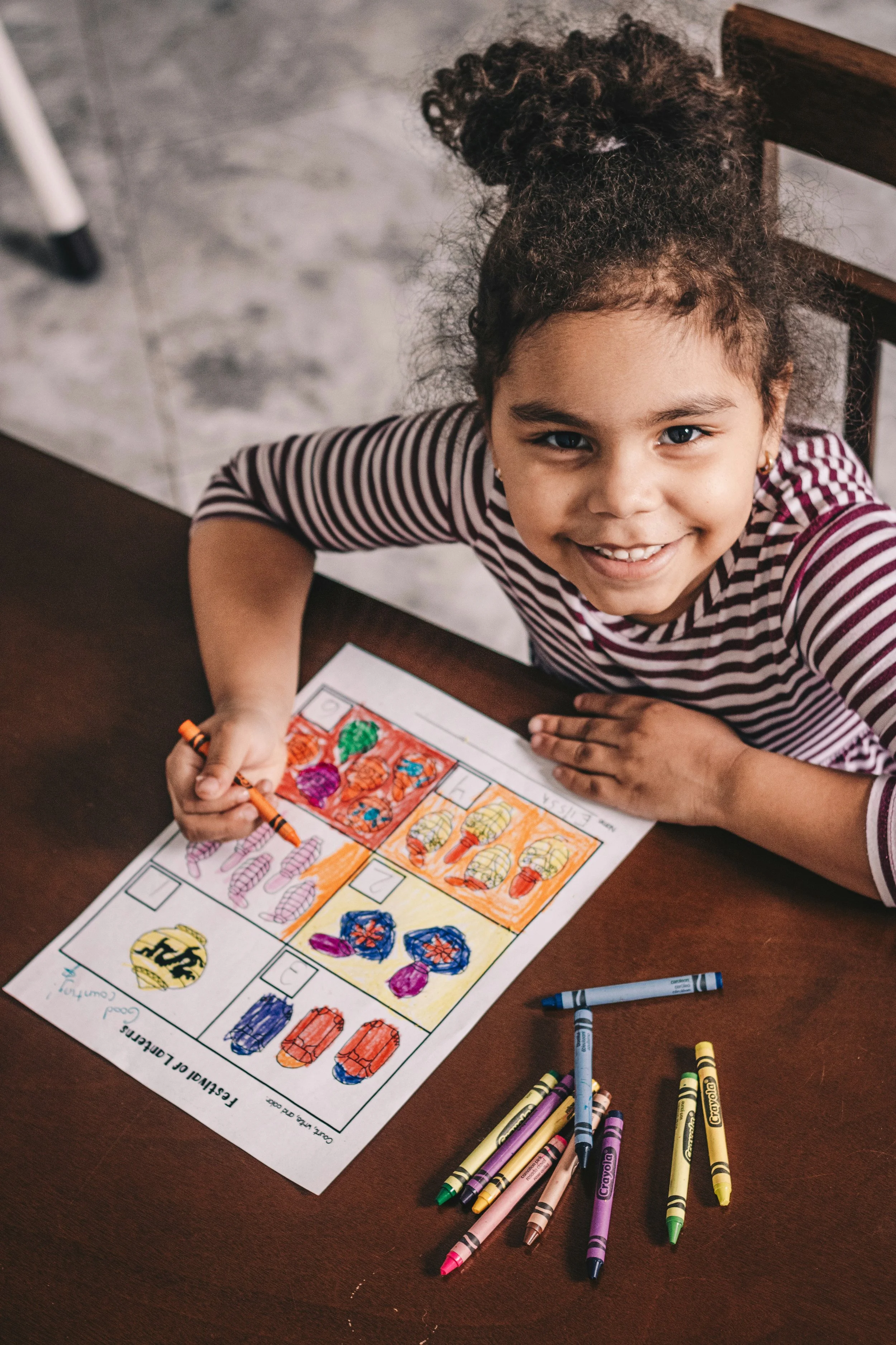 A young girl with curly hair pulled into a high bun, smiling, coloring a patriotic worksheet with markers. The worksheet features images of American flags, the Statue of Liberty, and other patriotic symbols, divided into sections with colorful backgr