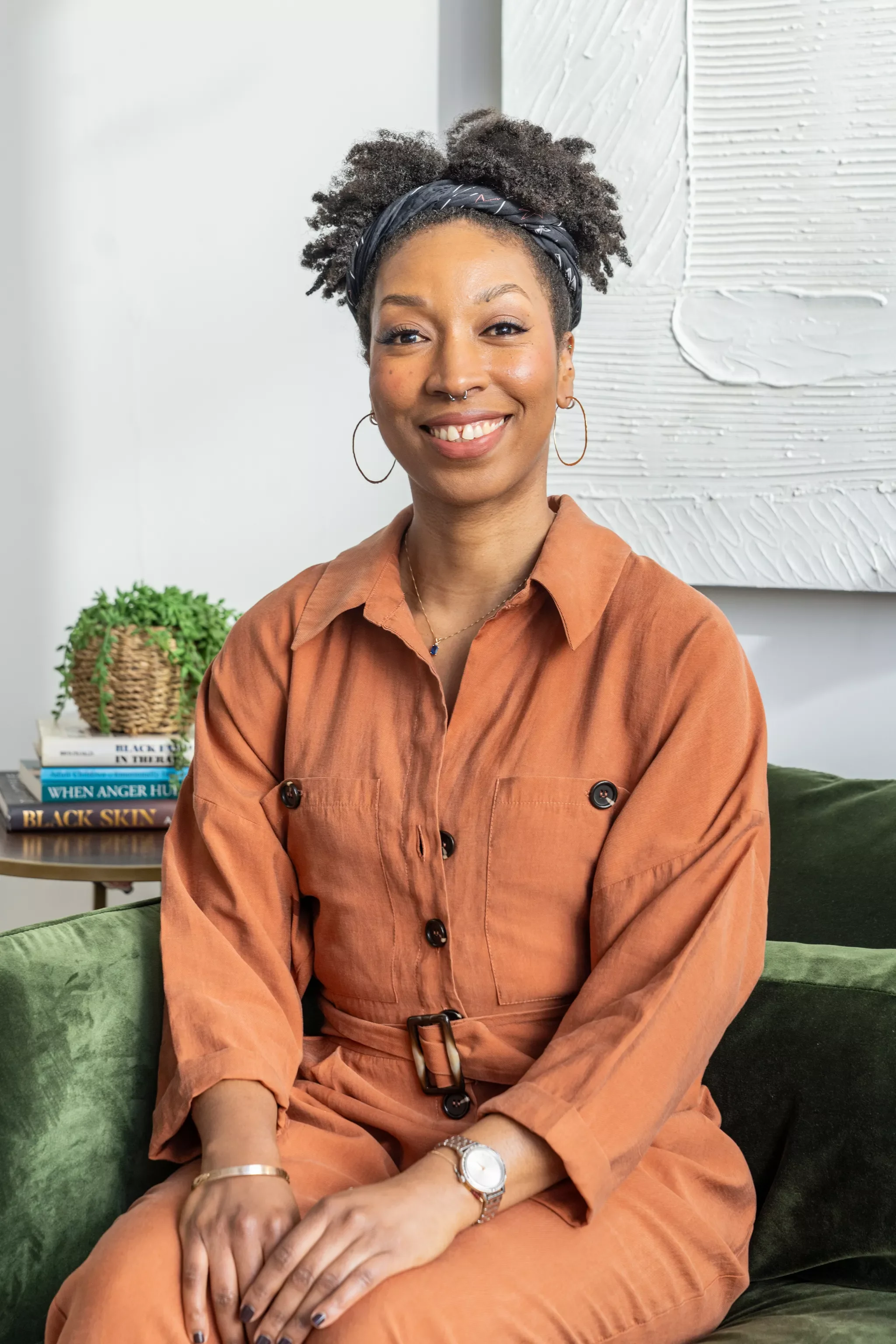 Portrait of a smiling woman with short curly hair, wearing a dark blue shirt, gold hoop earrings, a watch, and a bracelet, crossing her arms, standing in front of an abstract colorful painting.