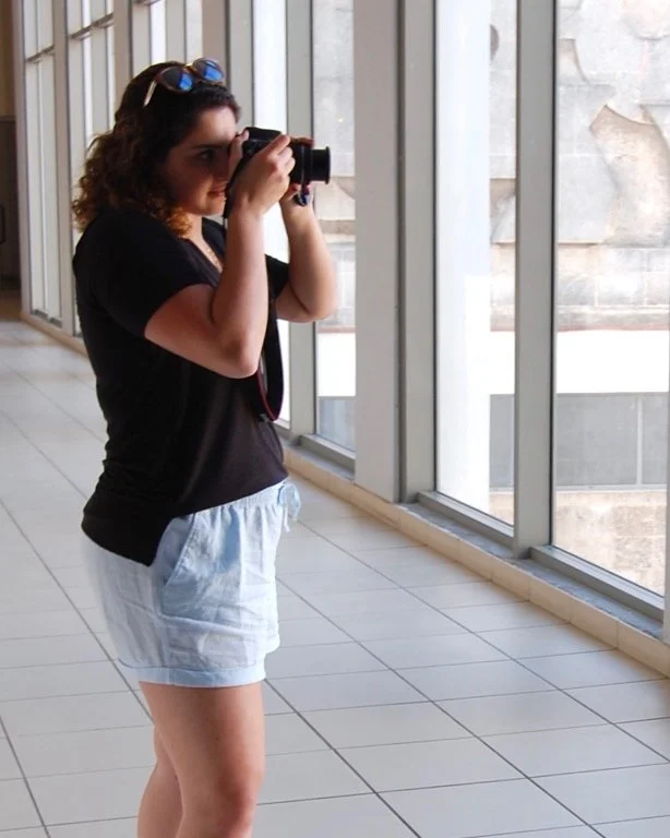 A woman with curly hair, wearing a black t-shirt and light blue shorts, is standing inside by large windows, taking a photograph with a camera.