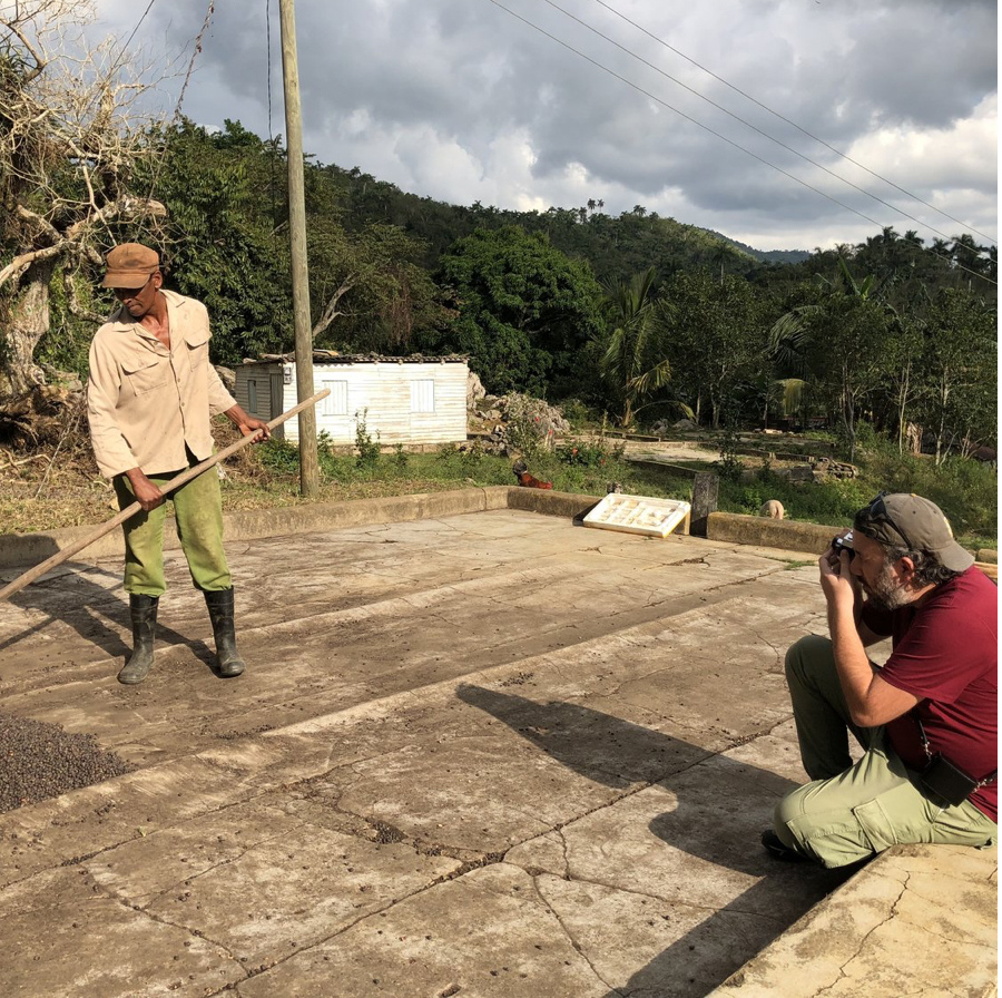 A man with a gray beard kneeling down, taking a photograph of another man who is using a rake on a cracked concrete surface outdoors in a rural area with green trees and mountains in the background.