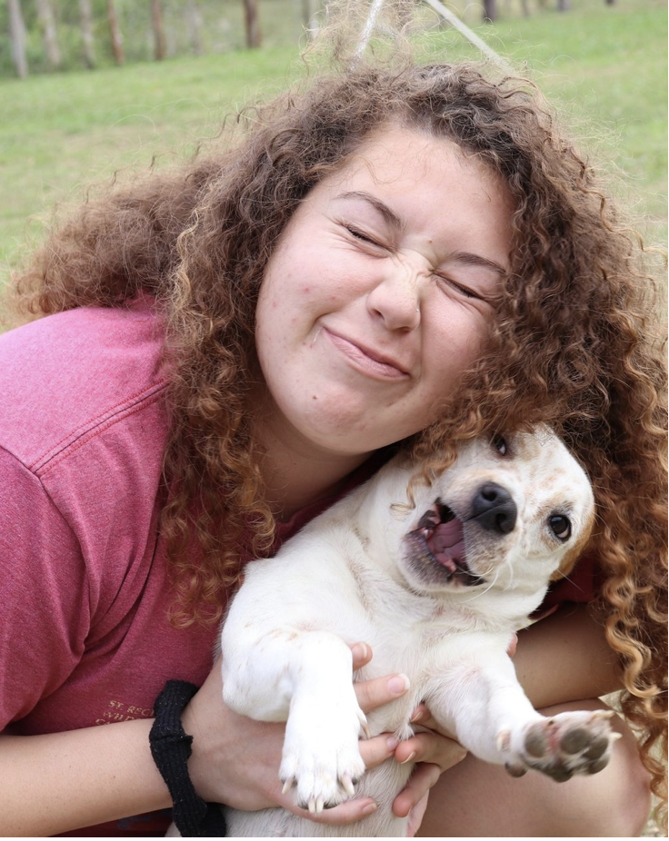 A girl with curly hair smiling and squinting joyfully while holding a small white puppy with brown spots outside on green grass.