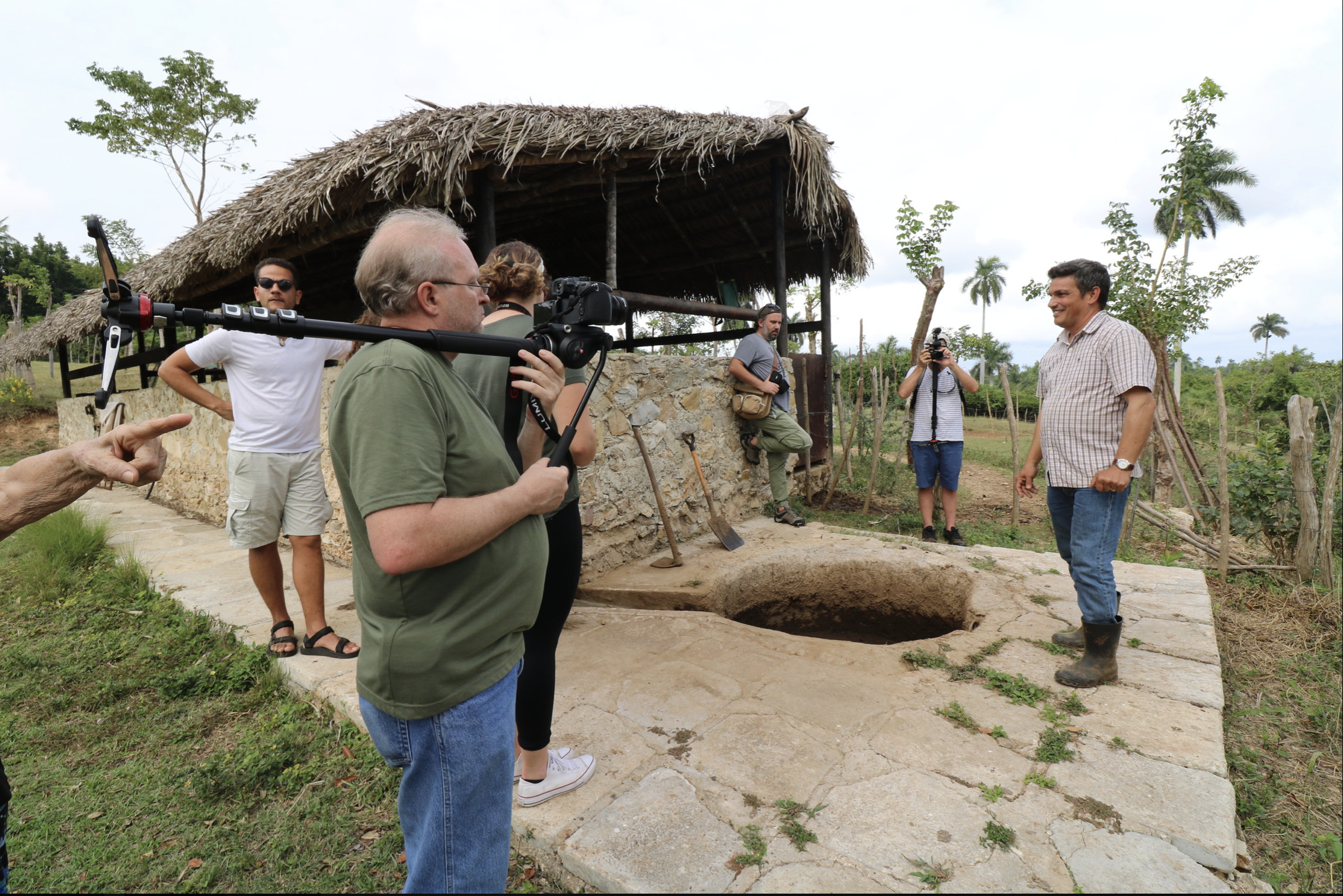 A group of people, some with cameras, visiting an archaeological site of a small Mayan pyramid with a step well, surrounded by green trees and grass, under a cloudy sky.