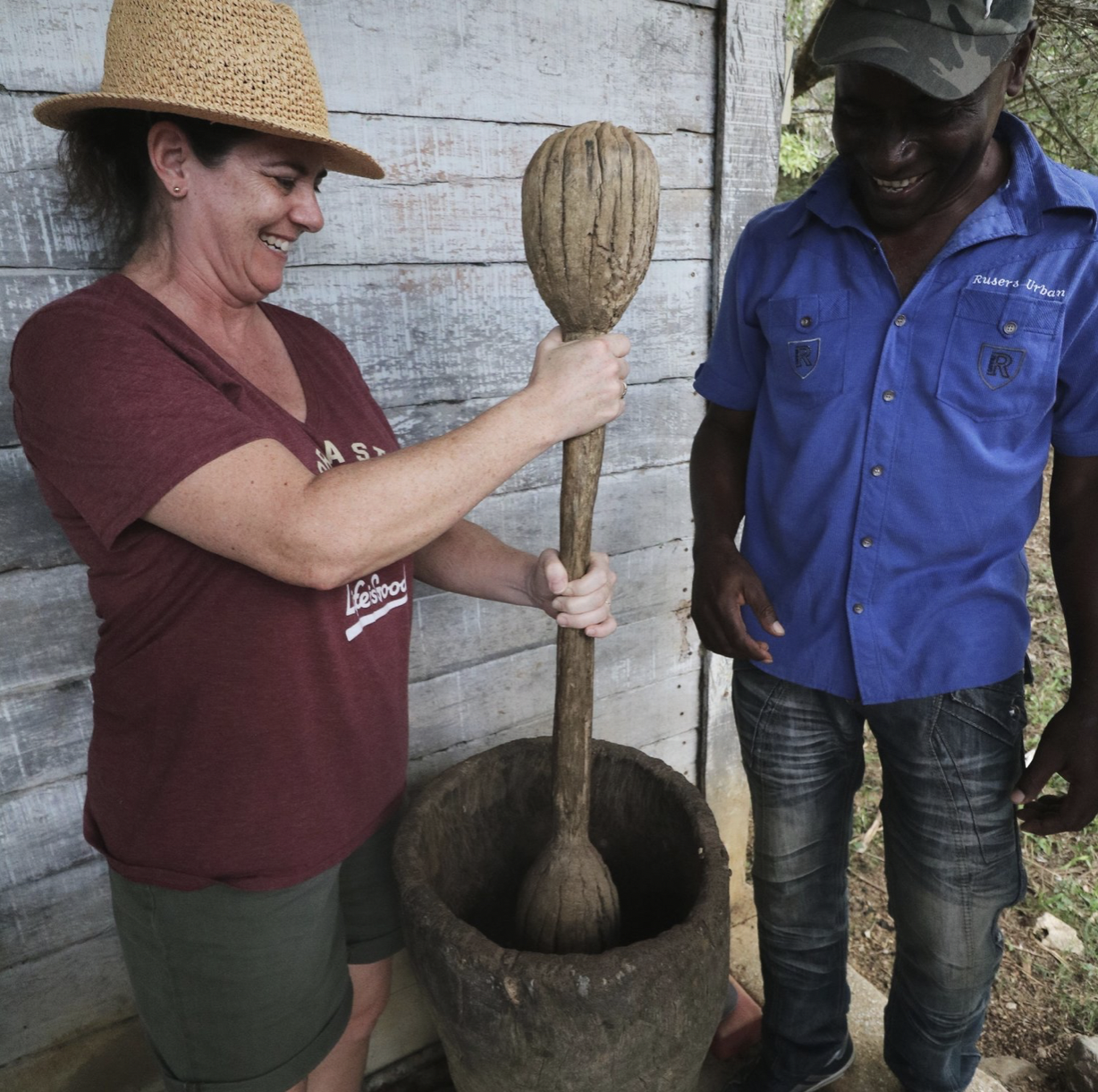 A woman and a man standing outdoors with a large wooden mortar and pestle, with the woman holding the pestle and the man smiling.