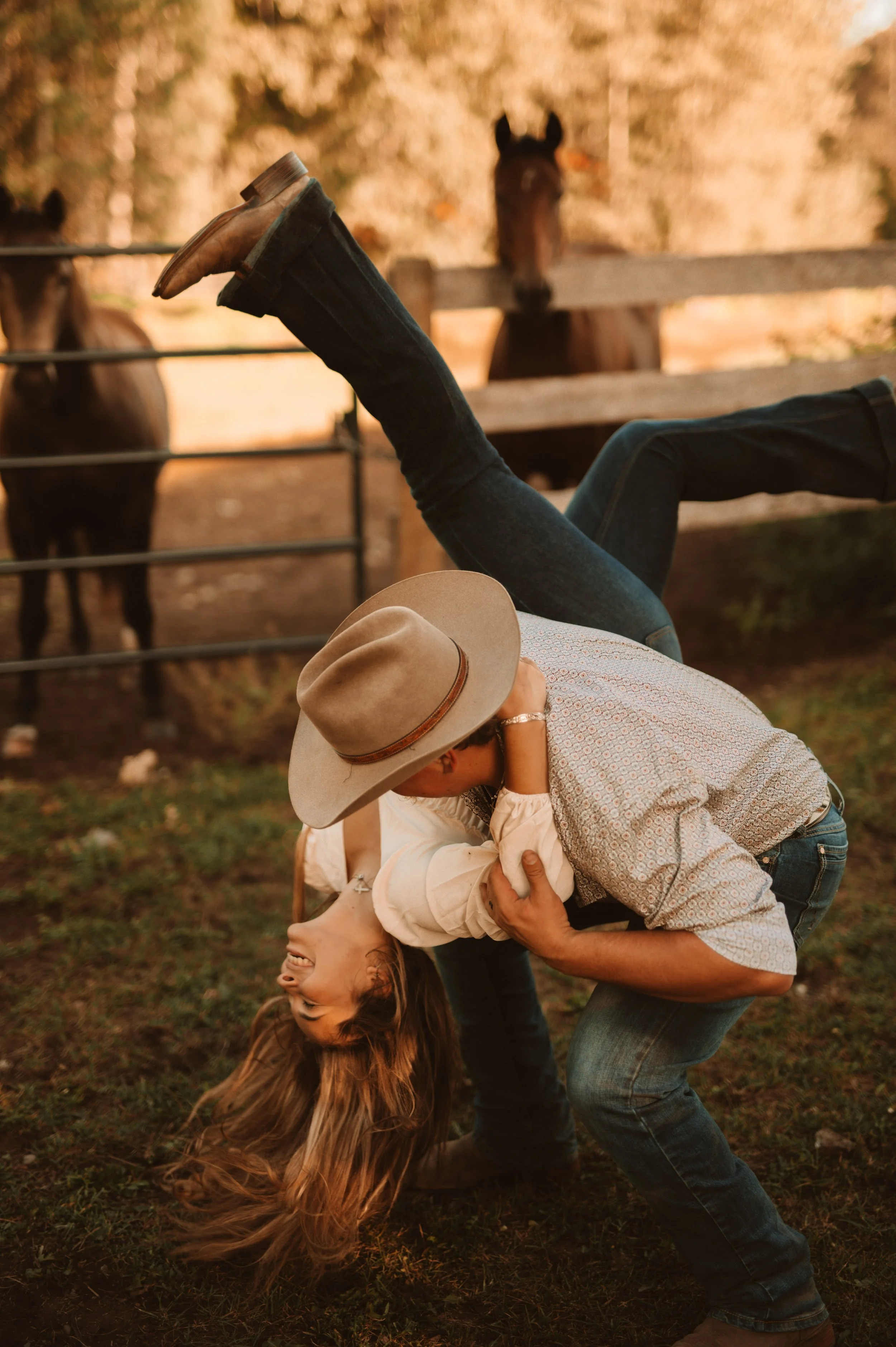 Two people engaged in a playful fight in a farm field with horses in the background, one wearing a hat and the other appearing to be laughing, during sunset.
