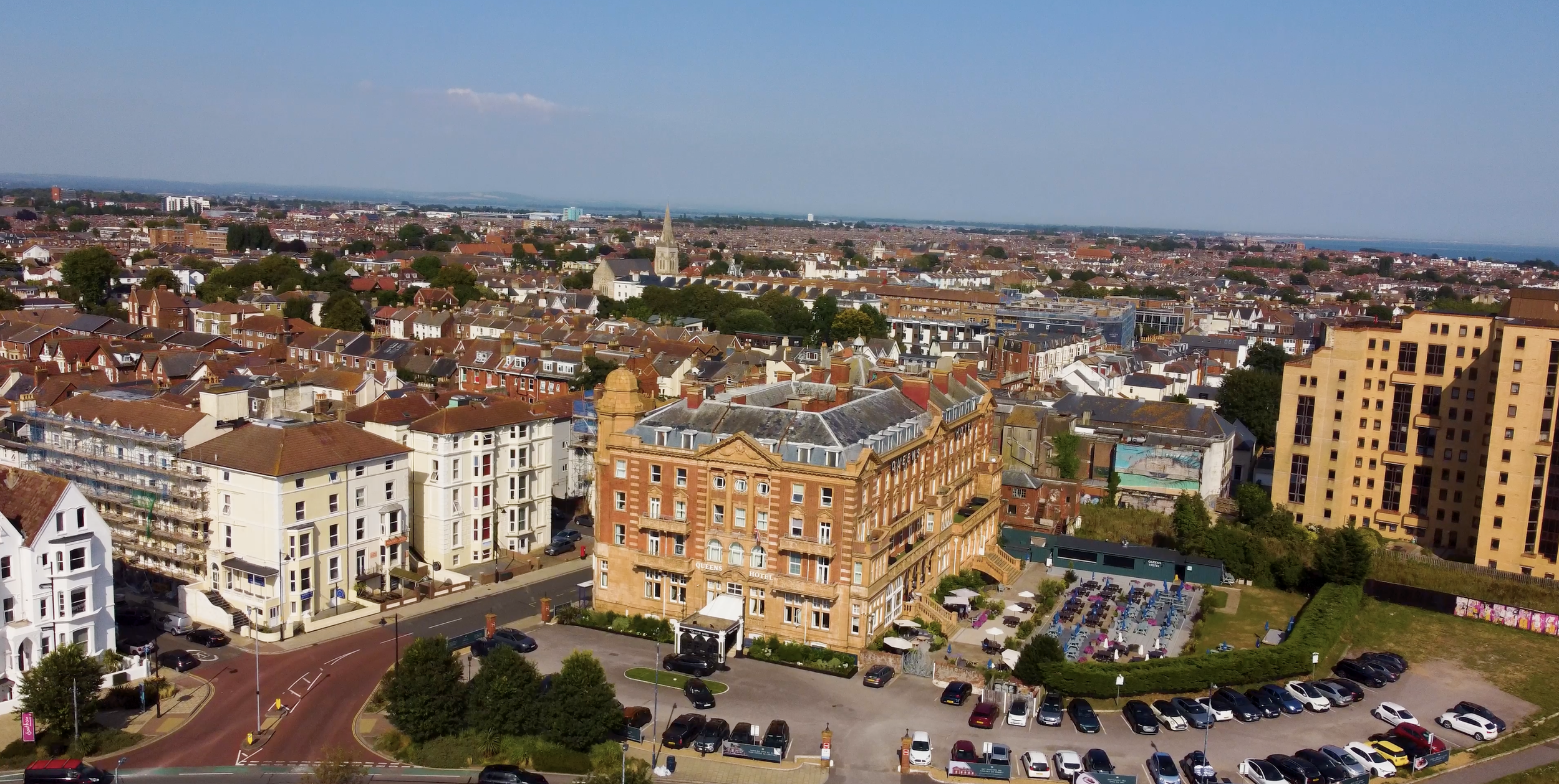 Aerial view of a city with a mix of historic and modern buildings, including a large hotel, residential apartments, and a parking lot, with a church steeple in the background and a body of water visible in the distance.
