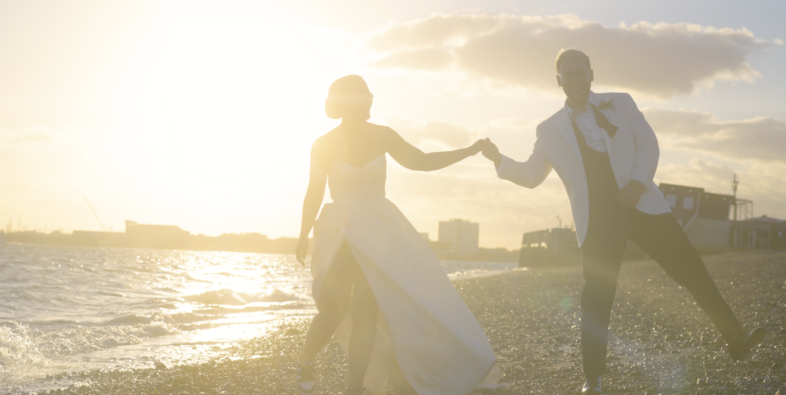 A couple in wedding attire holding hands and dancing on a beach during sunset.