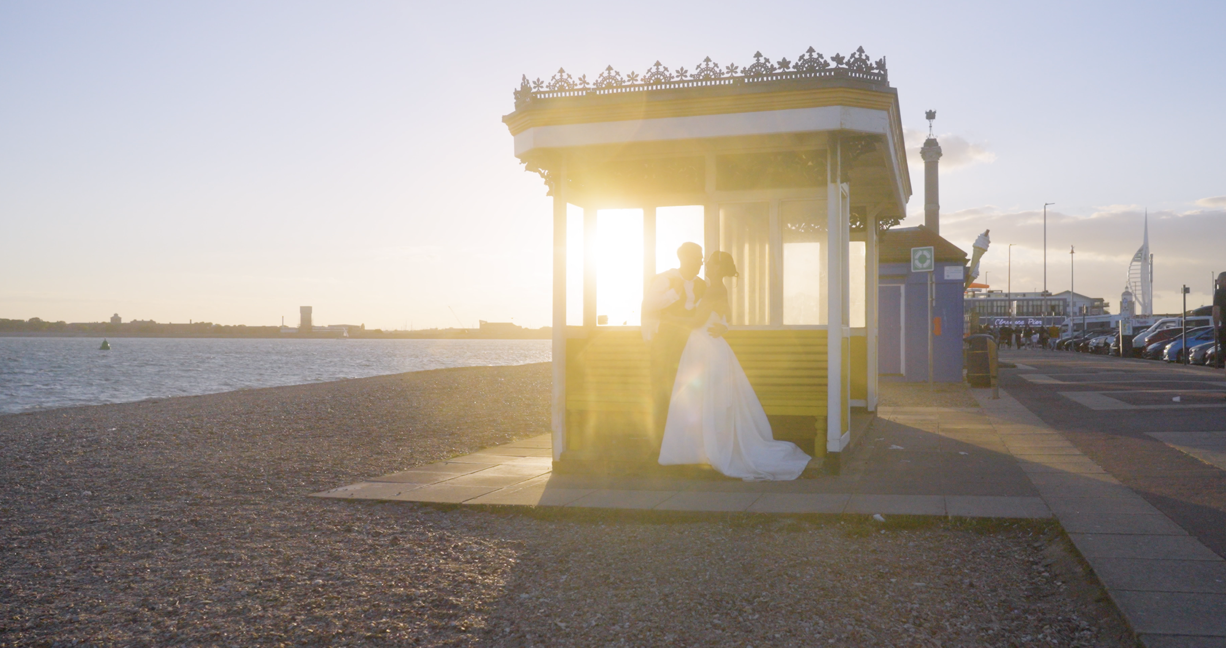 A bride and groom sharing a kiss at a beachside kiosk during sunset.