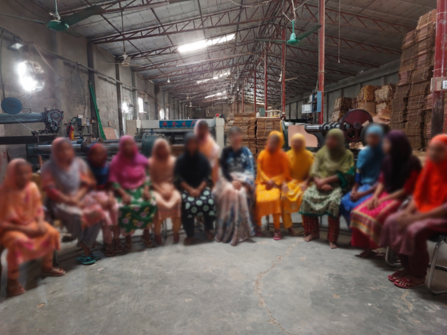 Group of women seated in a factory or warehouse surrounded by metal machinery and stacks of cardboard or paper on shelves.