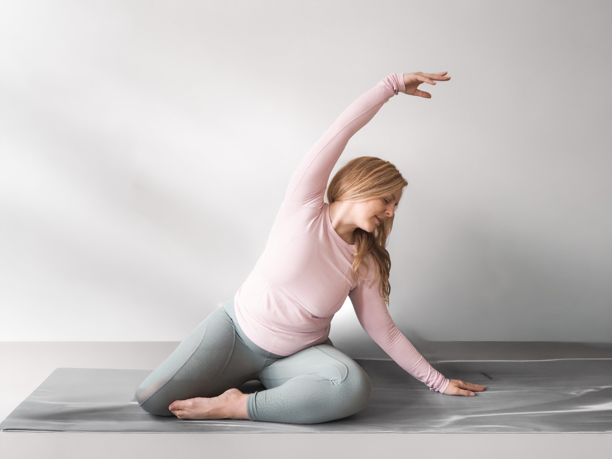 Somatic Pilates movement practice for midlife bone health and tension relief, showing an instructor demonstrating a fluid seated side bend on a gray mat.