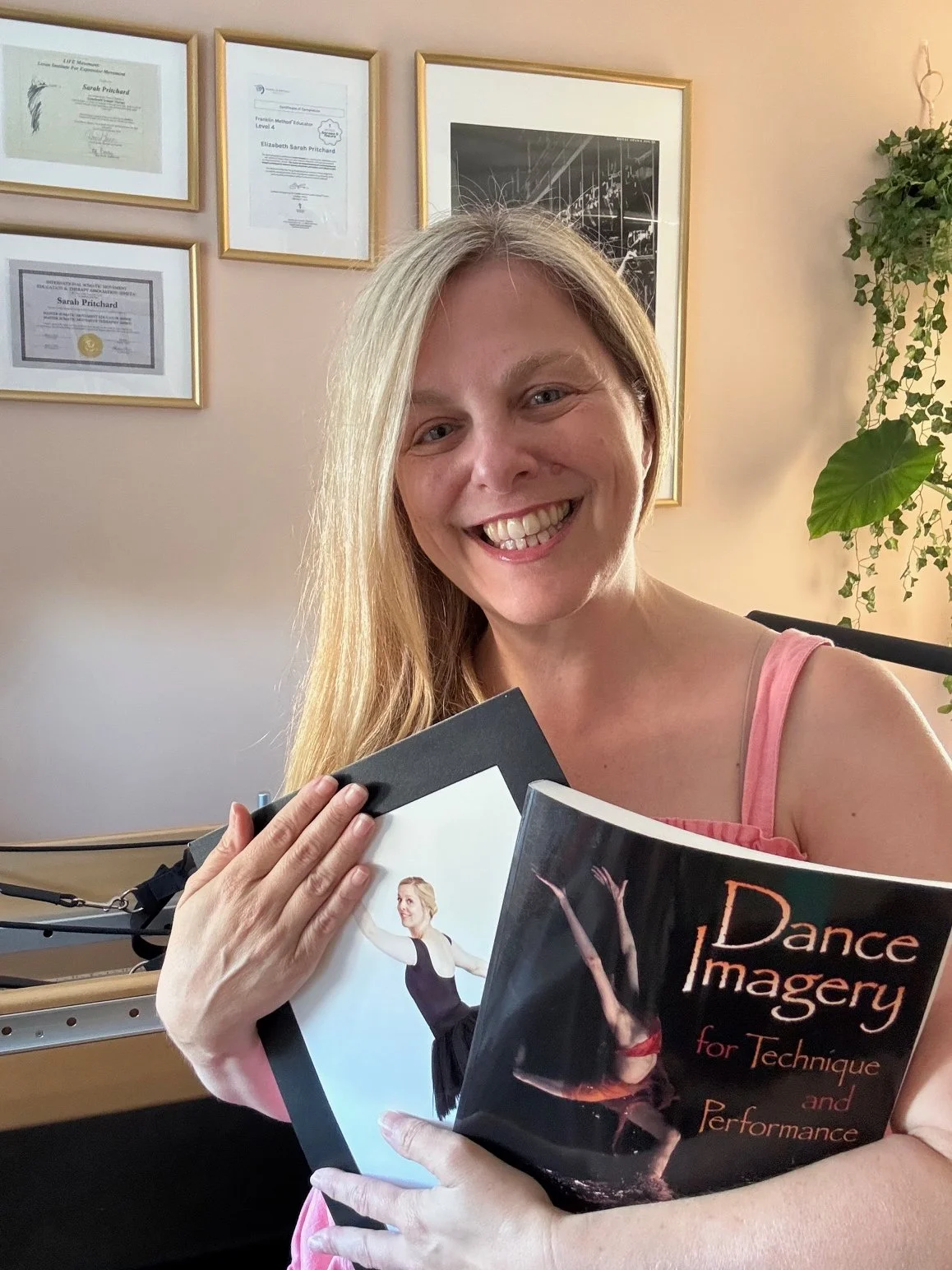 A portrait of Sarah Pritchard, MSMT/MSME, in her remedial Pilates and Somatic Movement studio, holding the book 'Dance Imagery' after guest teaching at the International Franklin Method® Studio. Behind her, framed accredited certificates are visible.