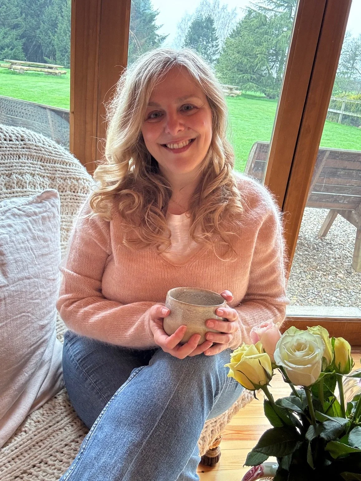 A trauma-informed SomaSoul somatic therapist sitting comfortably with a warm drink, offering a welcoming space for women seeking nervous system support