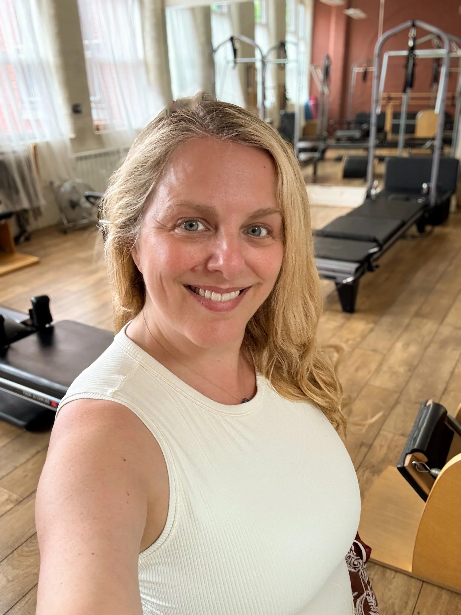 Sarah Pritchard, (MSMT/MSME) and 3rd Generation Pilates Specialist, smiling softly in her Jewellery Quarter studio in Birmingham. Classical studio equipment, including Pilates reformers and chairs, sits softly in the warm background.