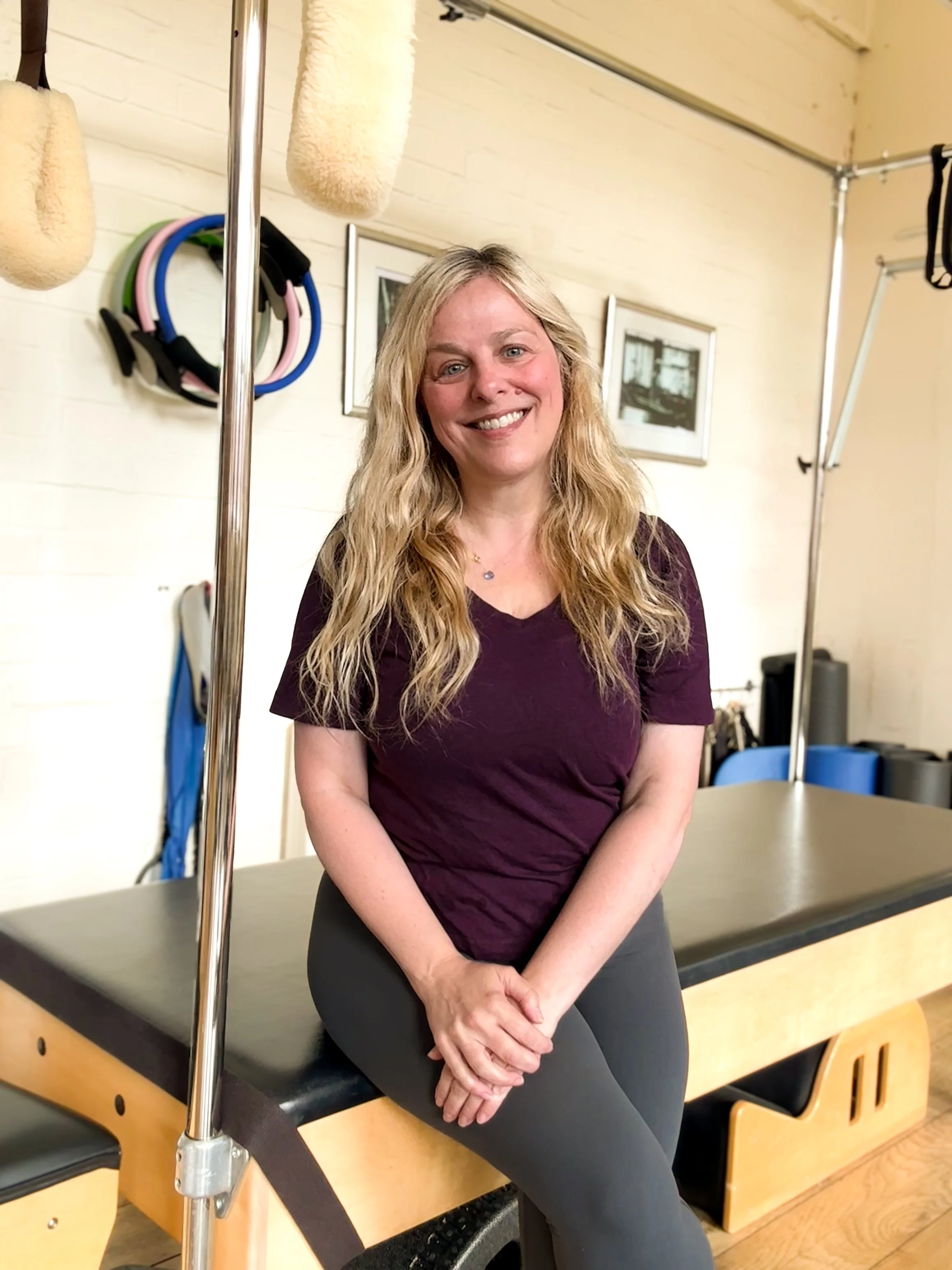 Sarah Pritchard smiling while sitting on a Pilates Cadillac machine in her sunny Birmingham Jewellery Quarter studio, representing specialist remedial Pilates and somatic movement.