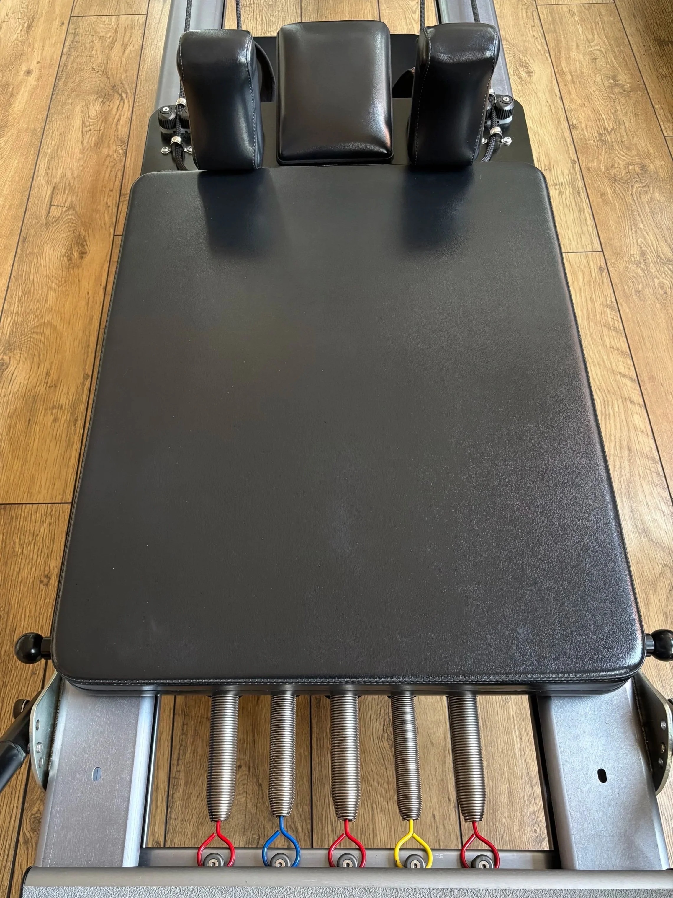 Top-down view of a modern Pilates Reformer machine in Sarah Pritchard's Birmingham Jewellery Quarter studio session, showing the black padded carriage, headrest, and five coloured resistance springs (red, blue, yellow) on a light wood floor.
