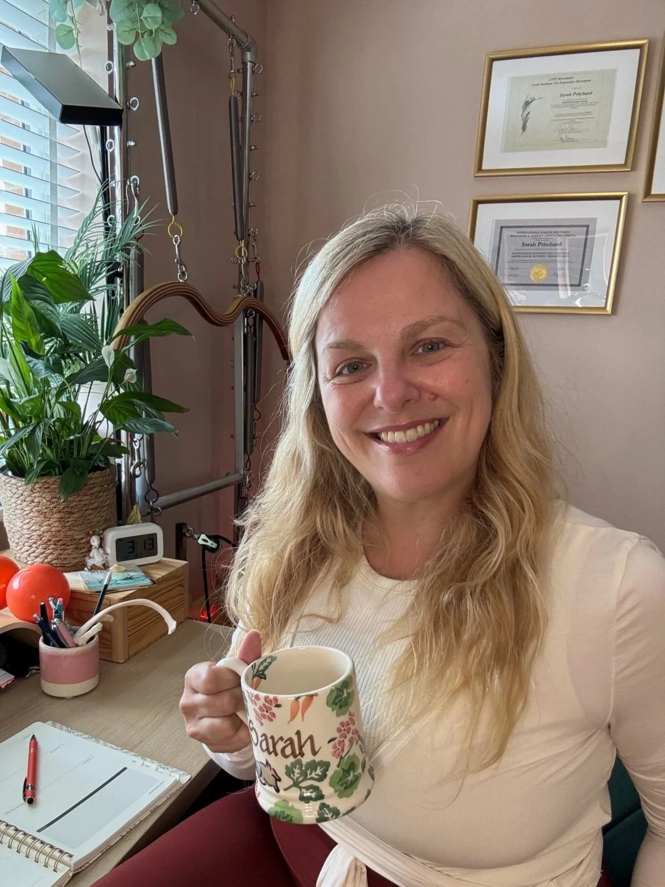 Sarah Pritchard smiling in her virtual Pilates studio at her desk, offering online Buff Bones and bone health movement sessions in Birmingham and throughout the UK.