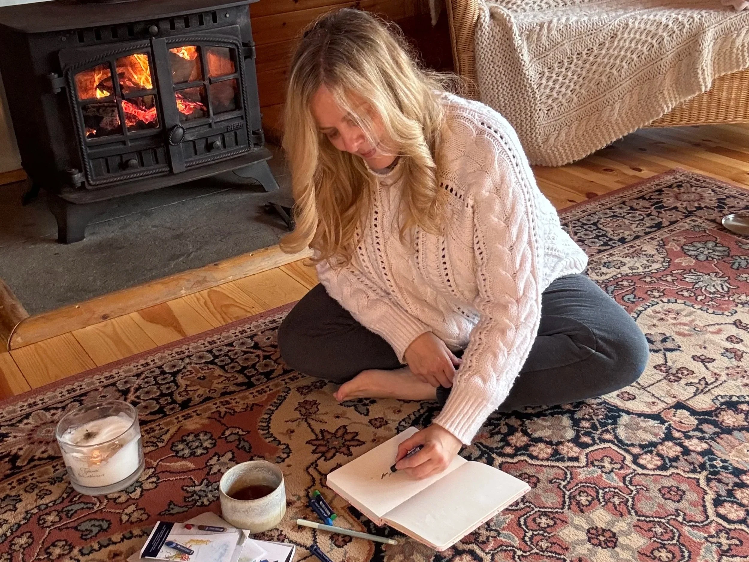 Sarah Pritchard engaging in the SomaSoul® expressive arts process by a warm fire, using therapeutic drawing as a somatic tool for emotional processing and integration.