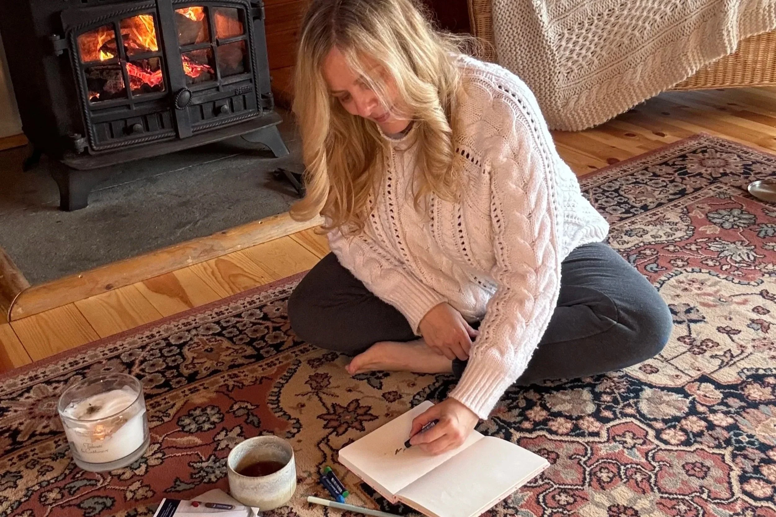Sarah Pritchard engaging in the SomaSoul® expressive arts process by a warm fire, using therapeutic drawing as a somatic tool for emotional processing and integration.