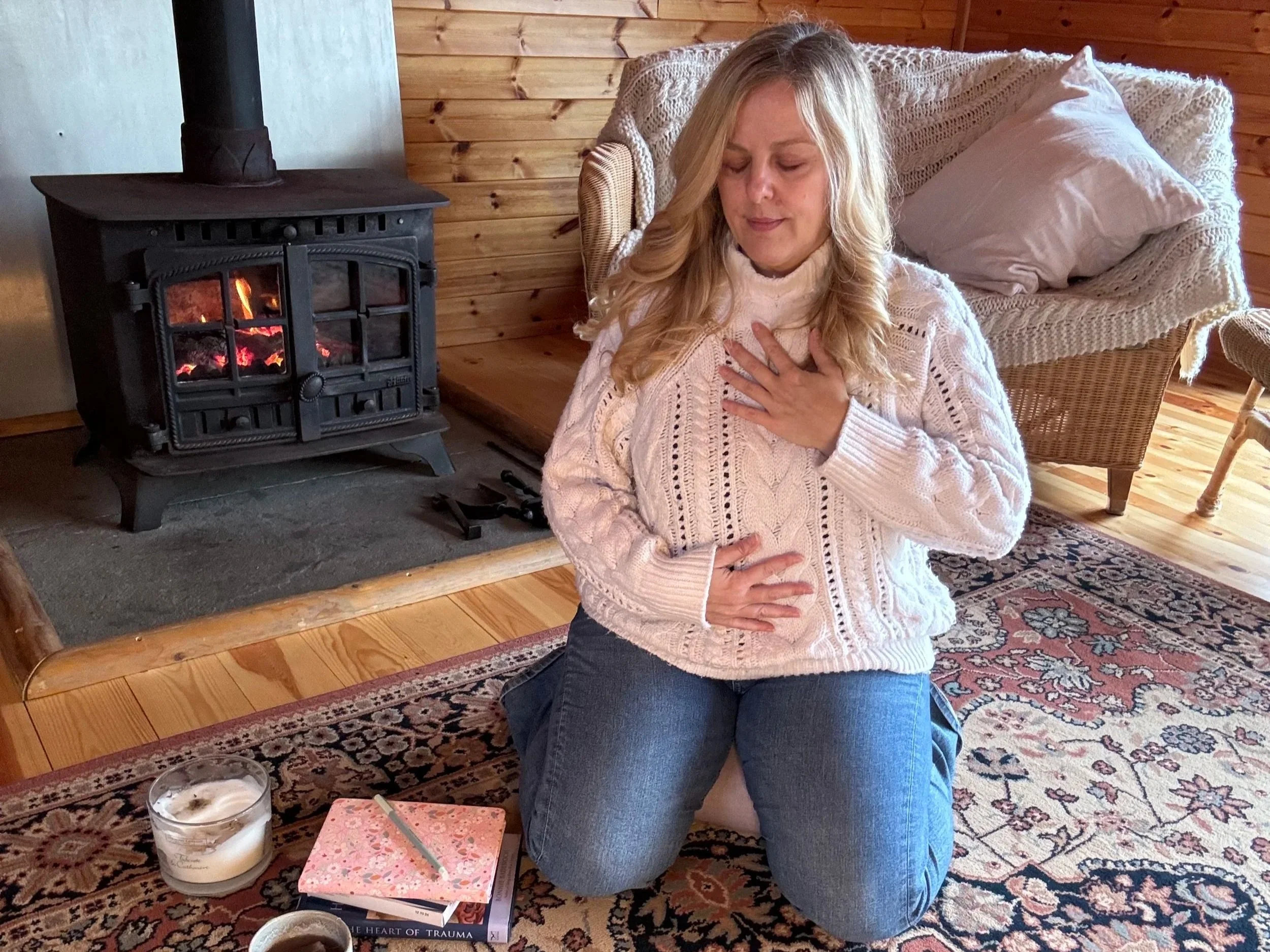 Sarah Pritchard, Somatic Therapist in a white jumper, practicing heart-centered somatic listening and nervous system regulation by a fire in her Shropshire studio.