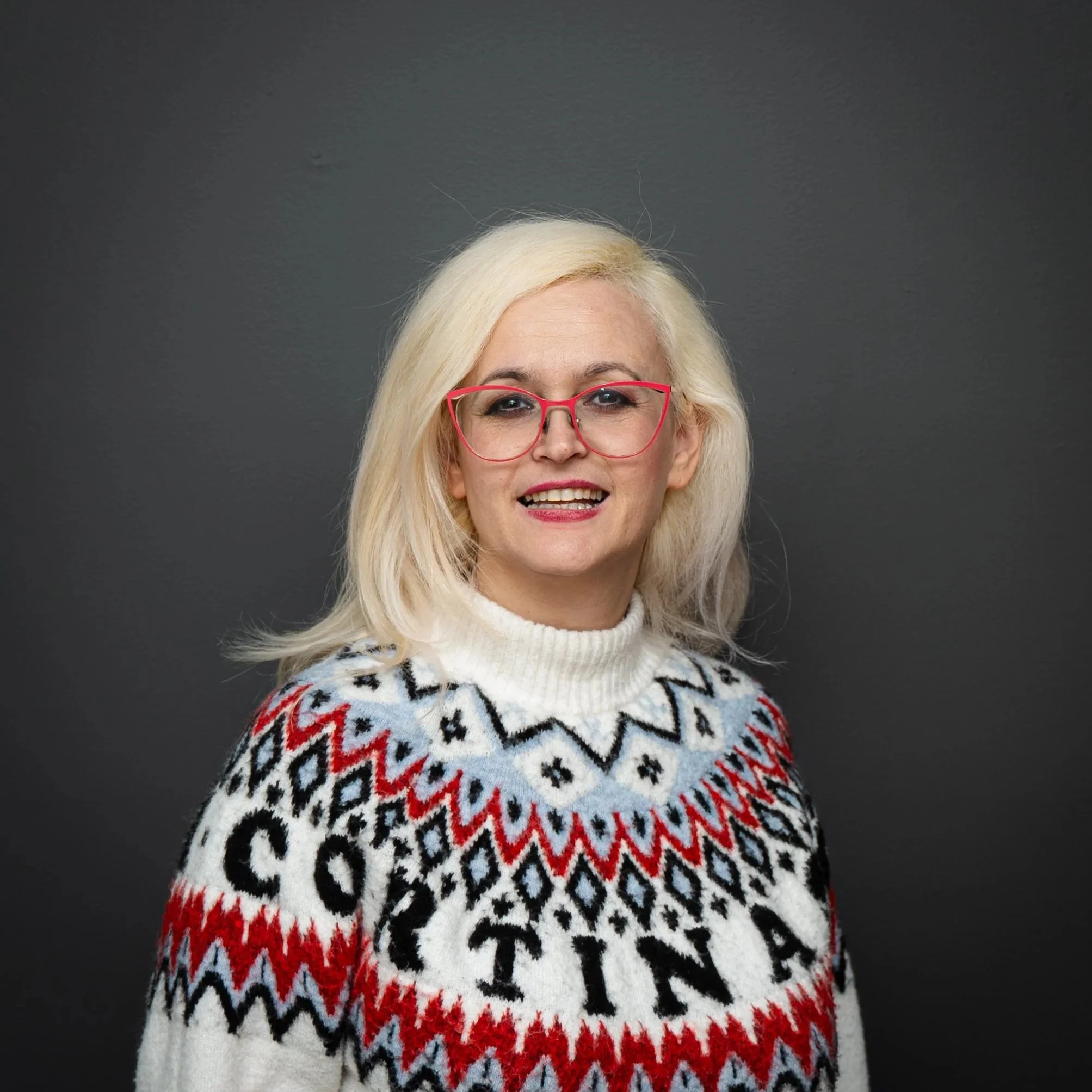 A woman with blonde hair wearing red glasses and a white sweater with Nordic patterns and the words 'SOCIETY' and 'ANX' against a dark grey background.