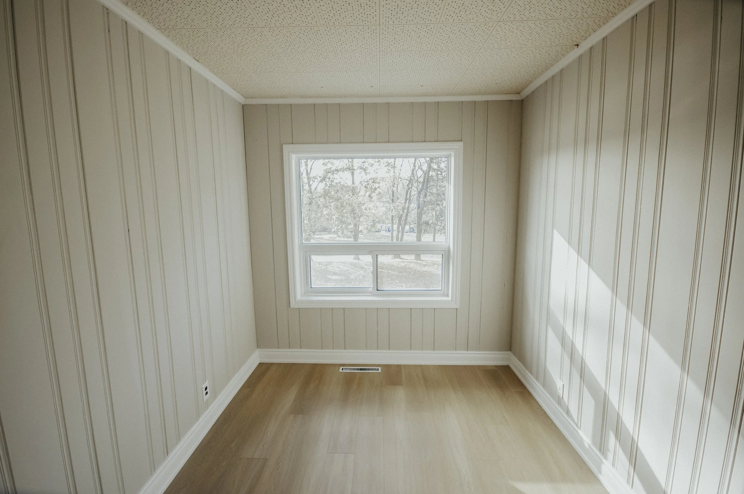 Empty room with light beige paneled walls, a wood floor, and a large window showing trees outside.