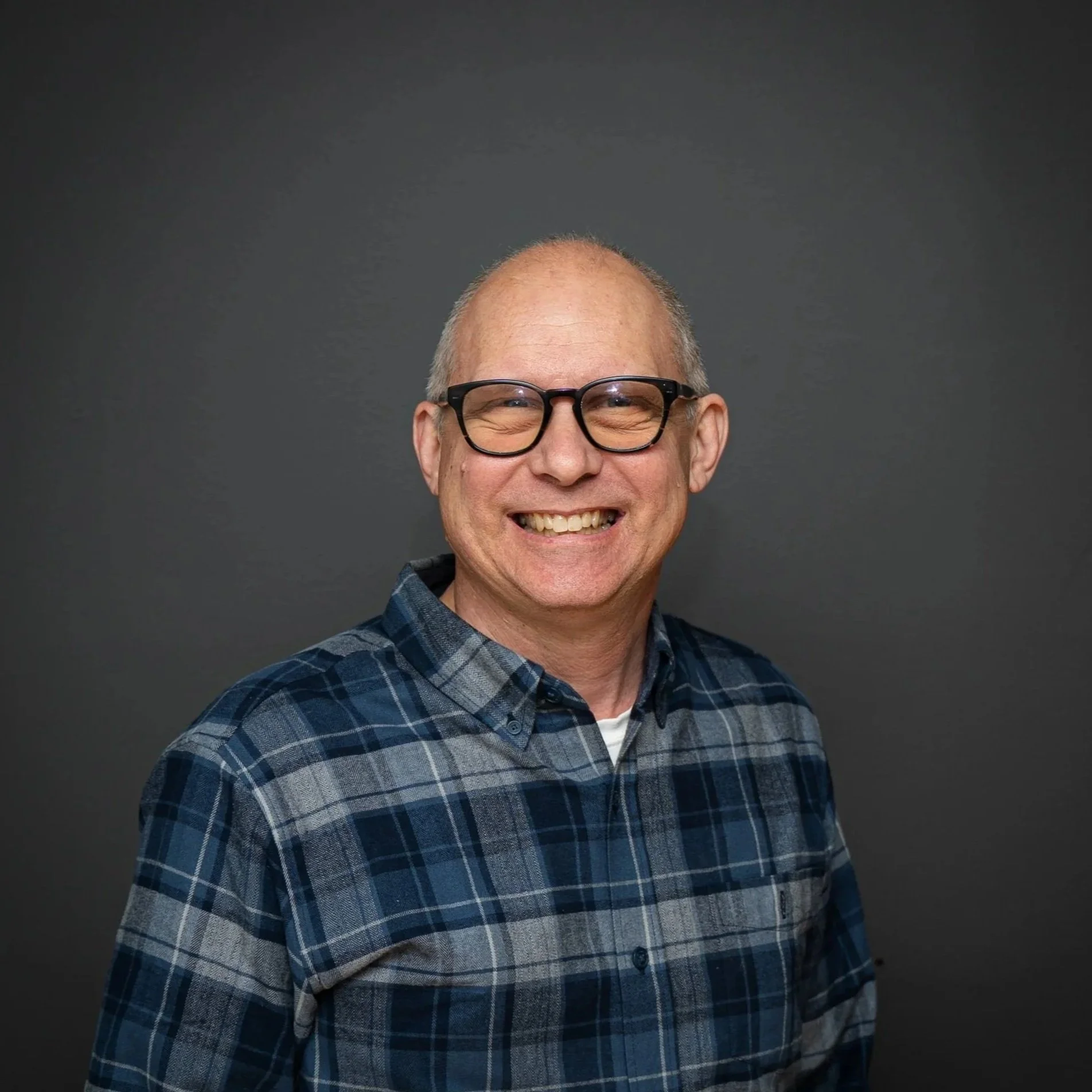 A middle-aged man with glasses and a big smile wearing a blue plaid shirt standing against a plain dark gray background.