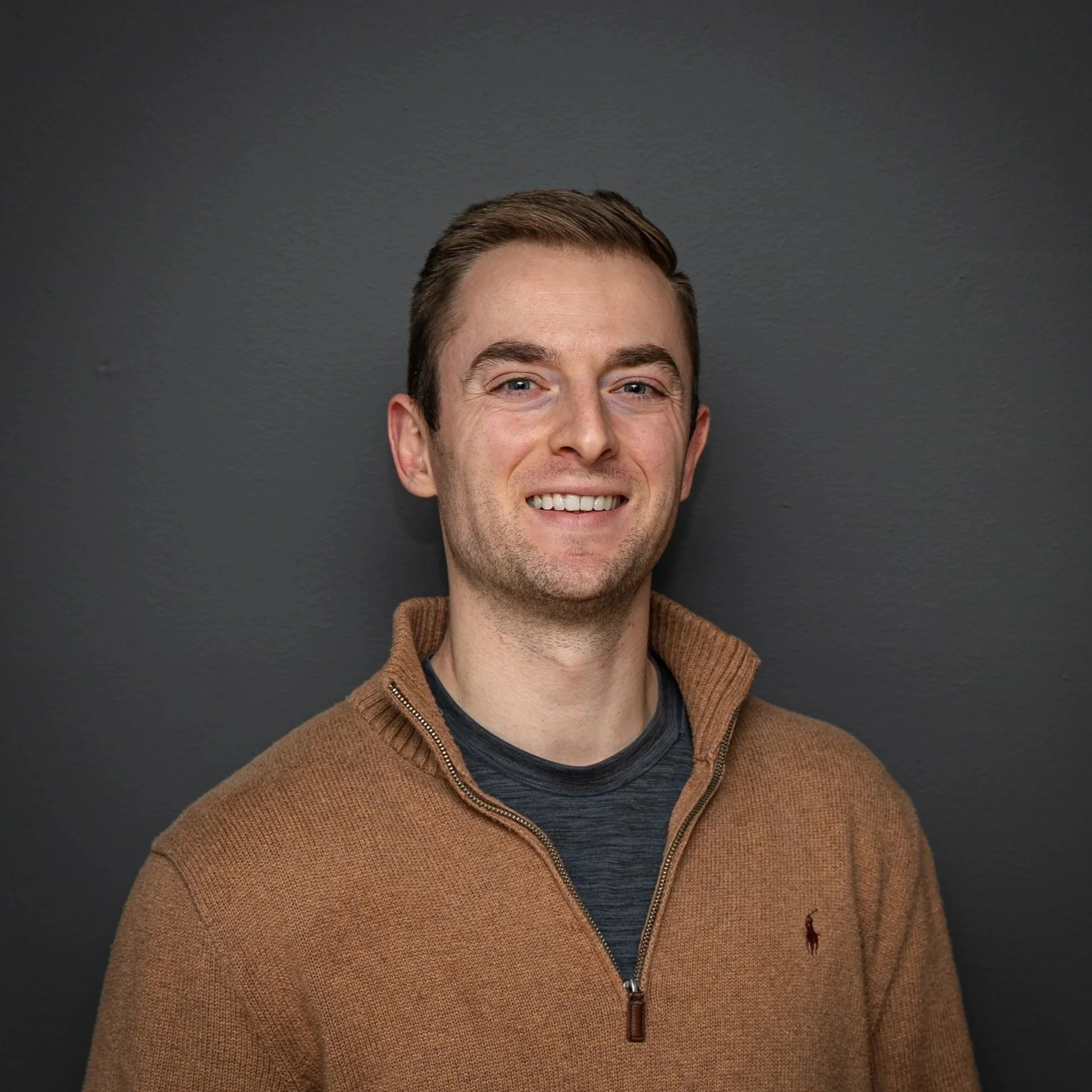 Portrait of a young man with short brown hair, blue eyes, smiling, wearing a brown zip-up sweater over a dark t-shirt, standing against a dark gray wall.