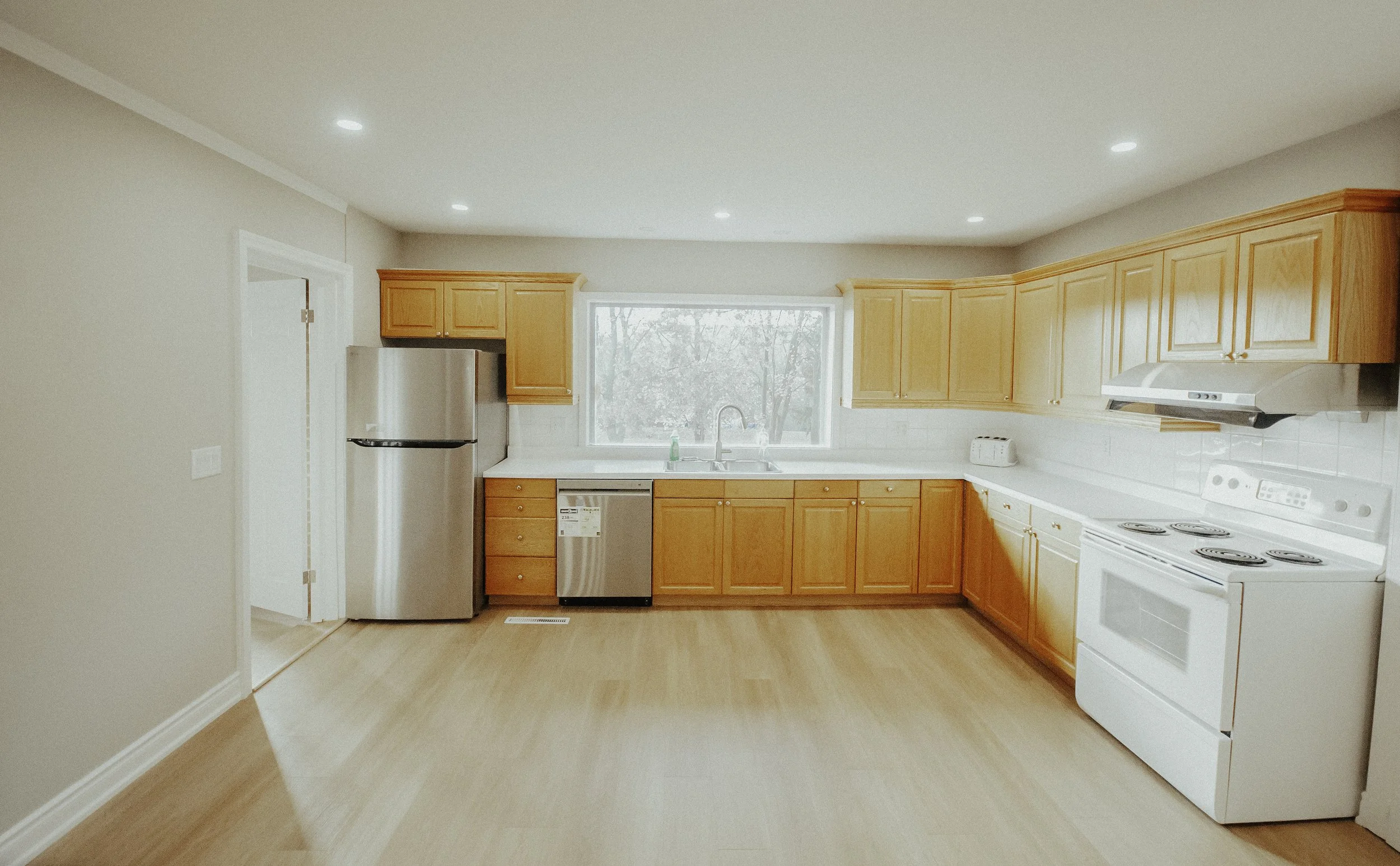 Empty kitchen with wooden cabinets, white countertops, stainless steel refrigerator, stove, and a window above the sink.