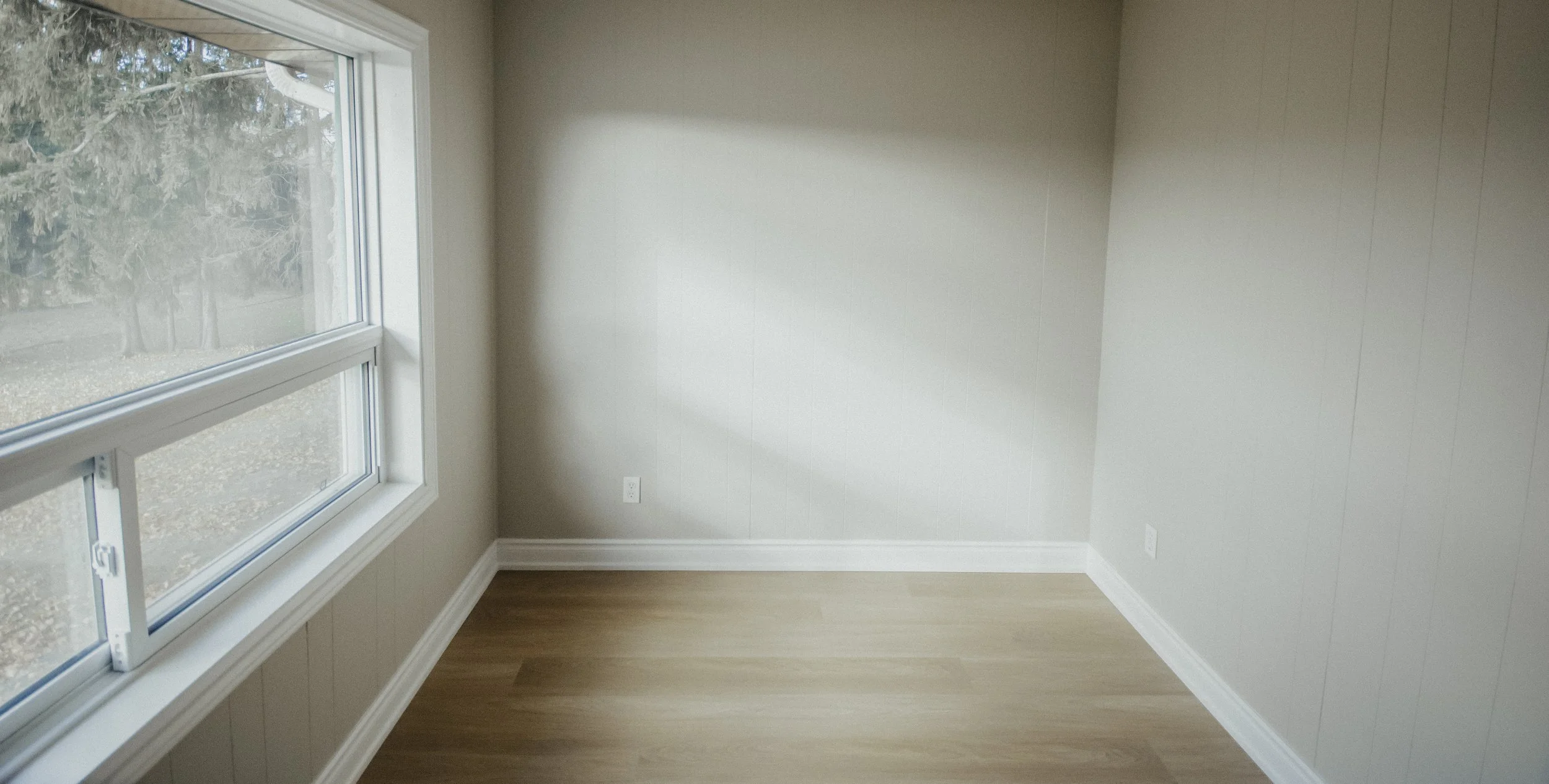 Empty room with beige walls, light wood flooring, and a large window showing trees outside.